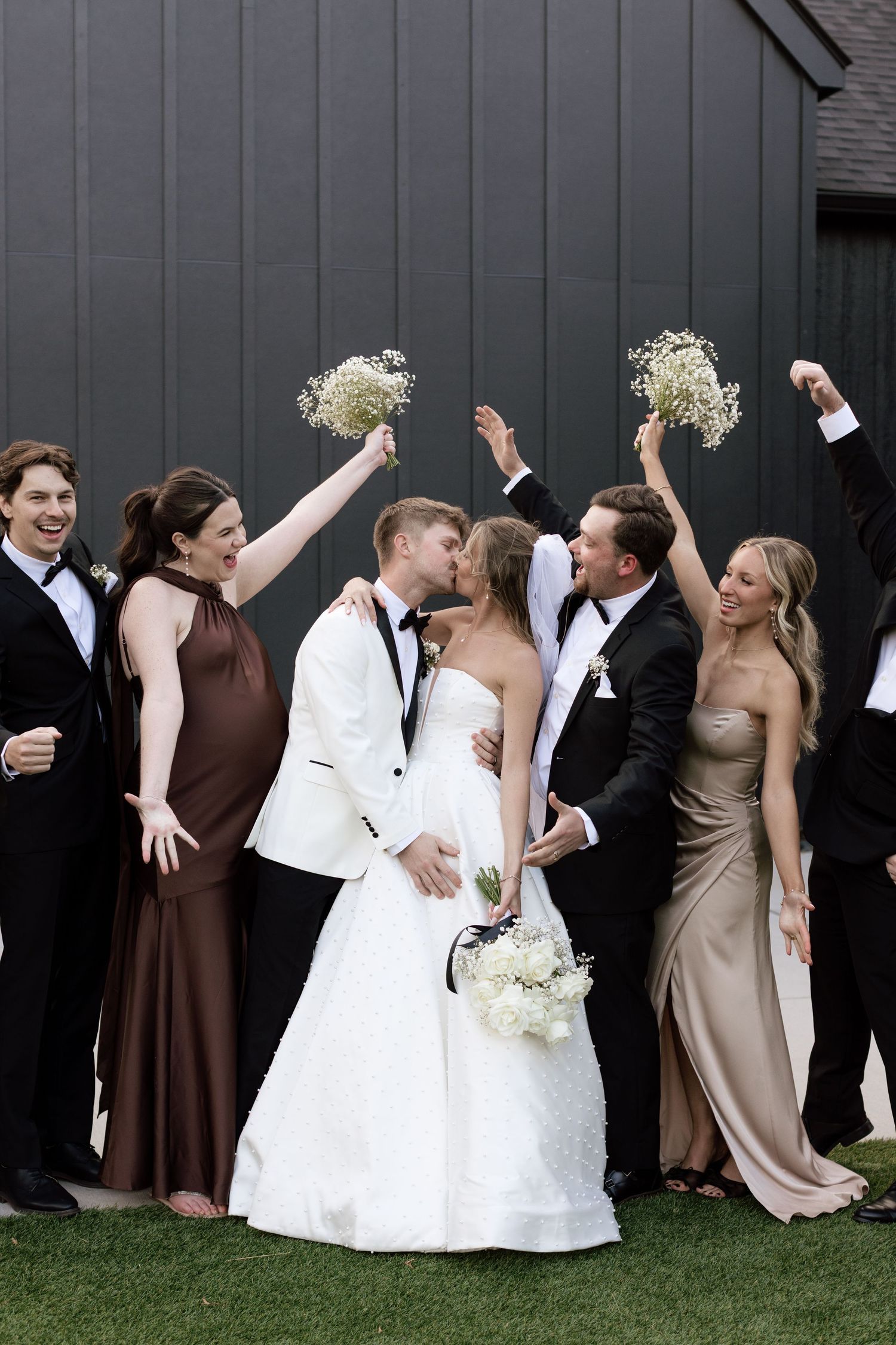 A bride and groom share a kiss surrounded by cheering bridesmaids and groomsmen outside a black barn.