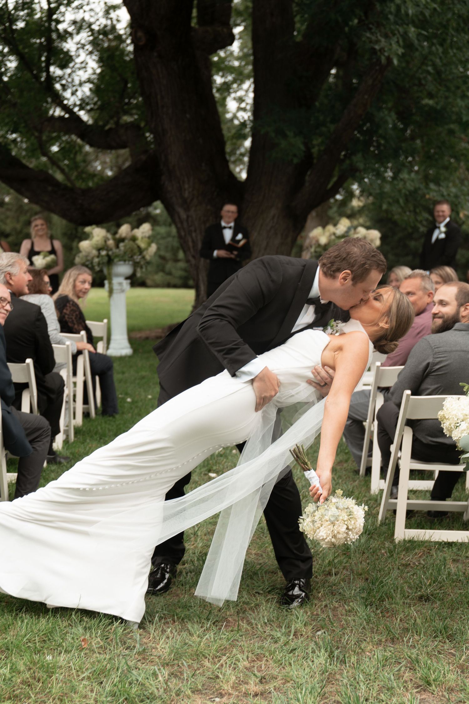 Wedding party poses for a creative falling domino-style photo on grass under large trees at an outdoor ceremony.