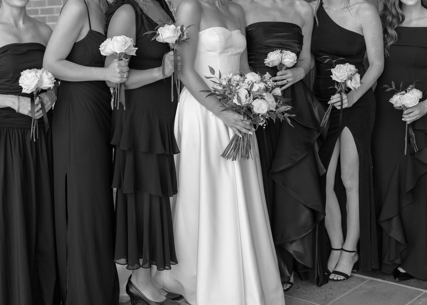 Bride and bridesmaids in black dresses stand together holding white floral bouquets at formal portraits.