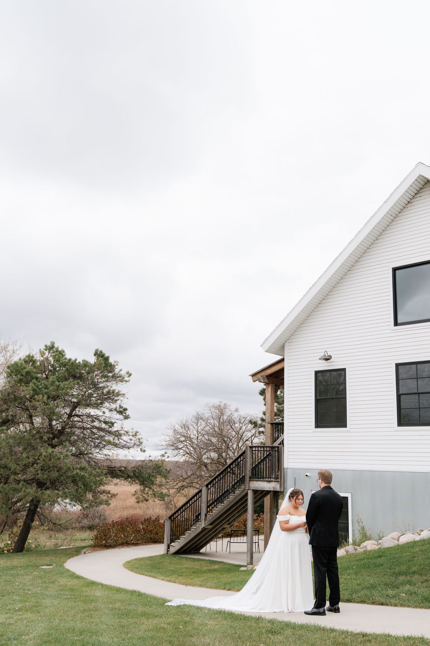 A couple in wedding attire stands beside a modern white building with exterior stairs on a cloudy day while they privately read their vows.