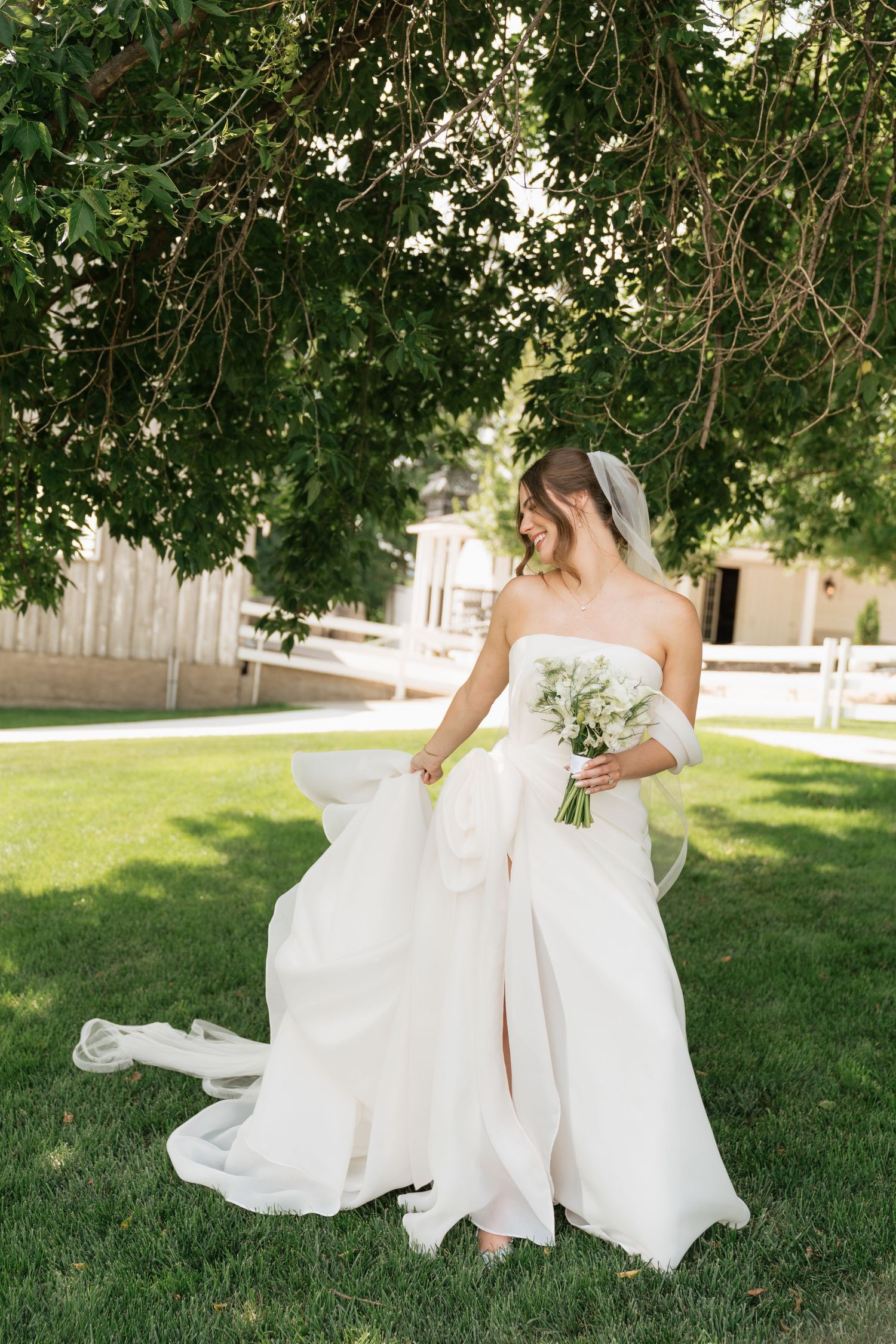 Bride in strapless white gown holding bouquet, standing on green lawn beneath lush tree canopy.