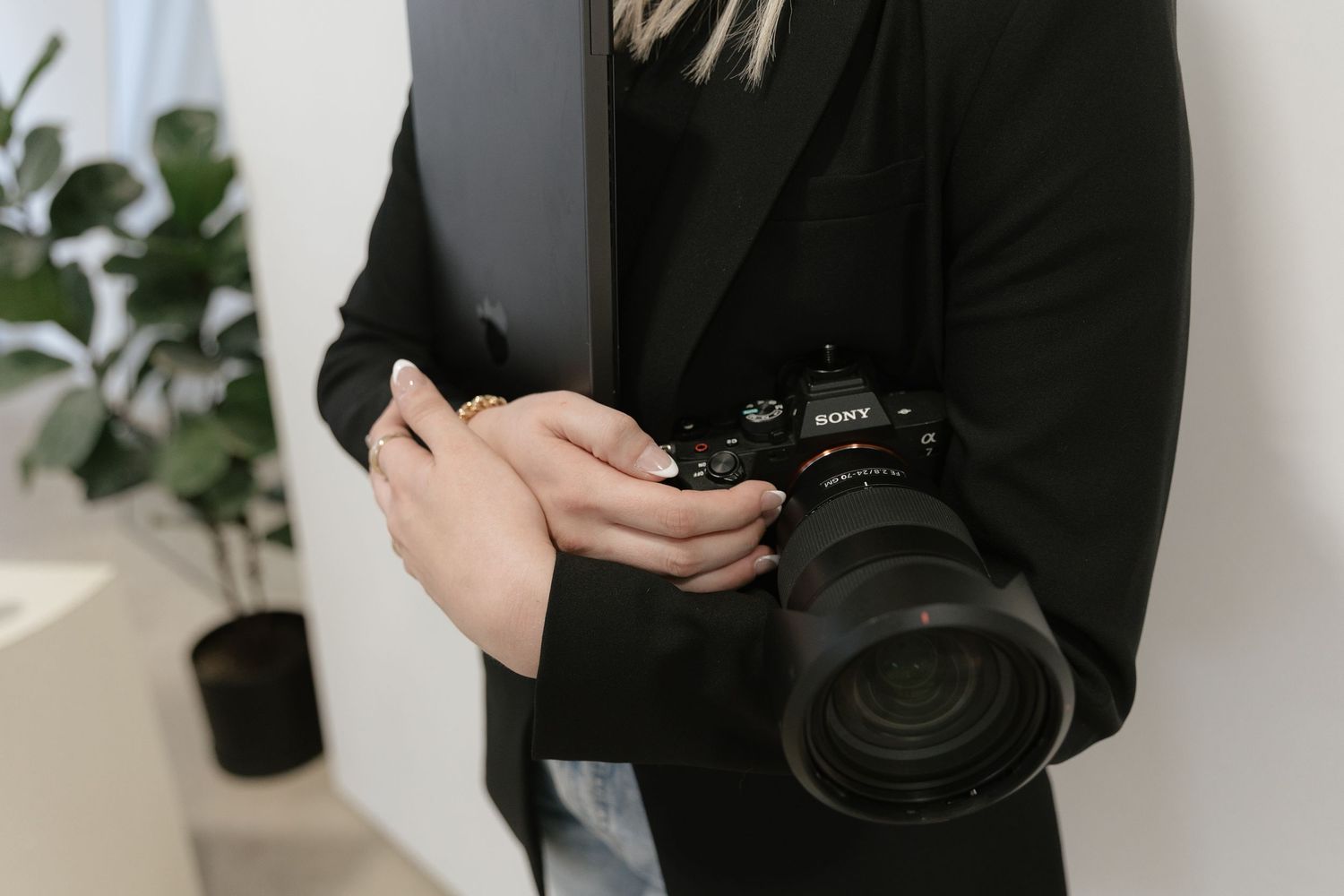 A photographer in black attire holds a camera while posing against a white wall with plants. Rochester, Minnesota wedding photographer.