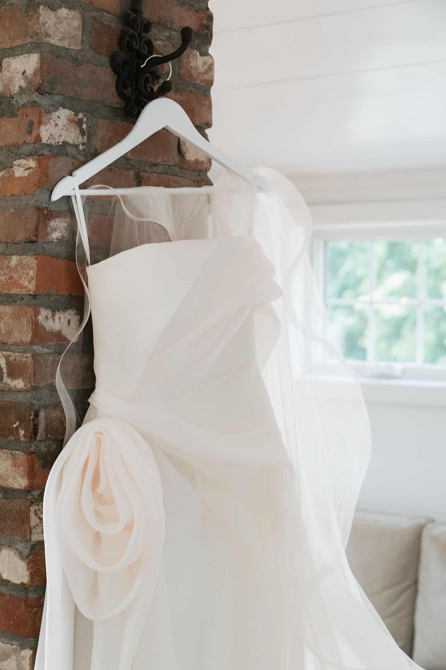 Elegant white wedding dress with floral detail hanging on a white hanger against a rustic brick wall near a window.