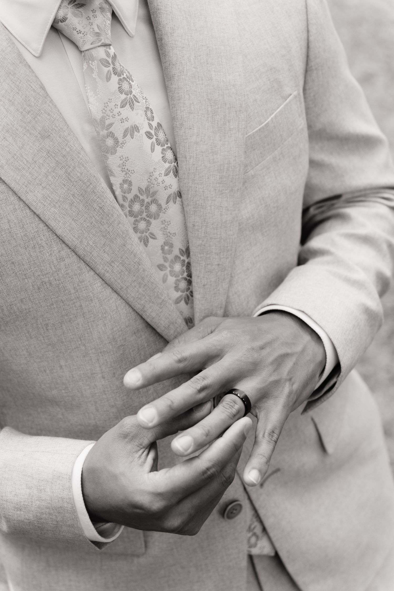 Black and white photo of two grooms holding hands, displaying wedding rings, wearing matching light suits with floral ties.