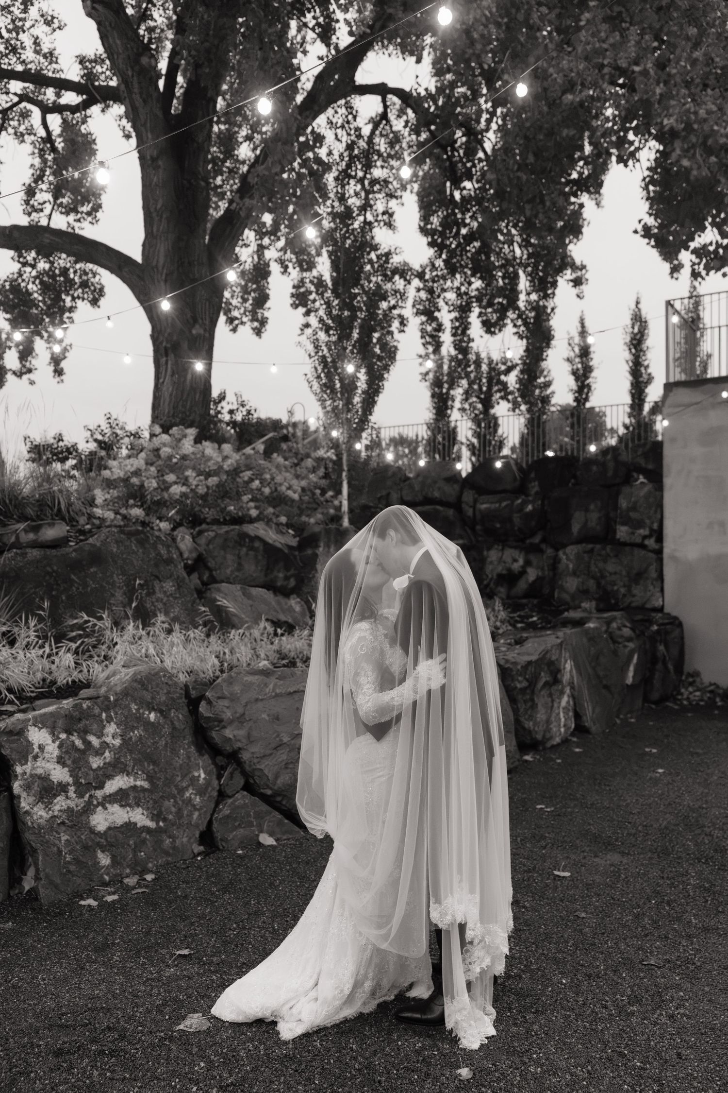 Black and white photo of bride and groom kissing under flowing veil outdoors with string lights and stone wall.