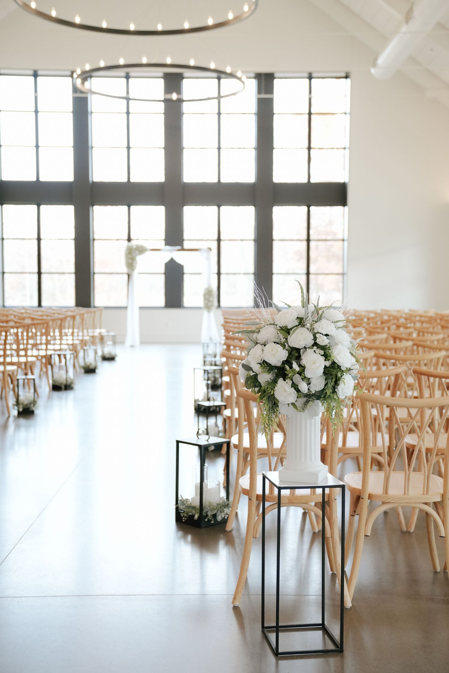 Modern white event space with wooden crossback chairs and elegant floral arrangements set up for a wedding ceremony.