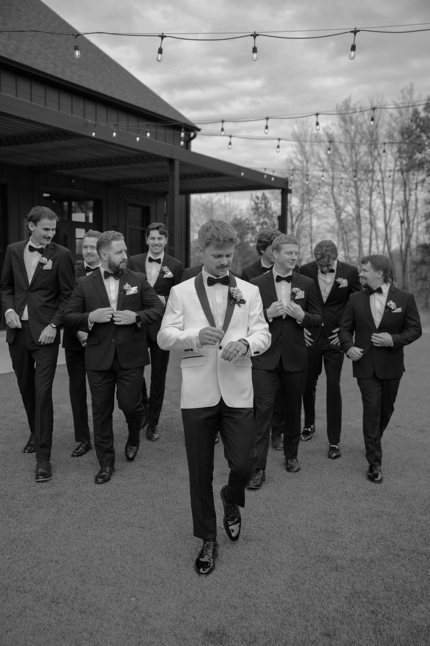 Groomsmen in black tuxedos running together across a lawn.