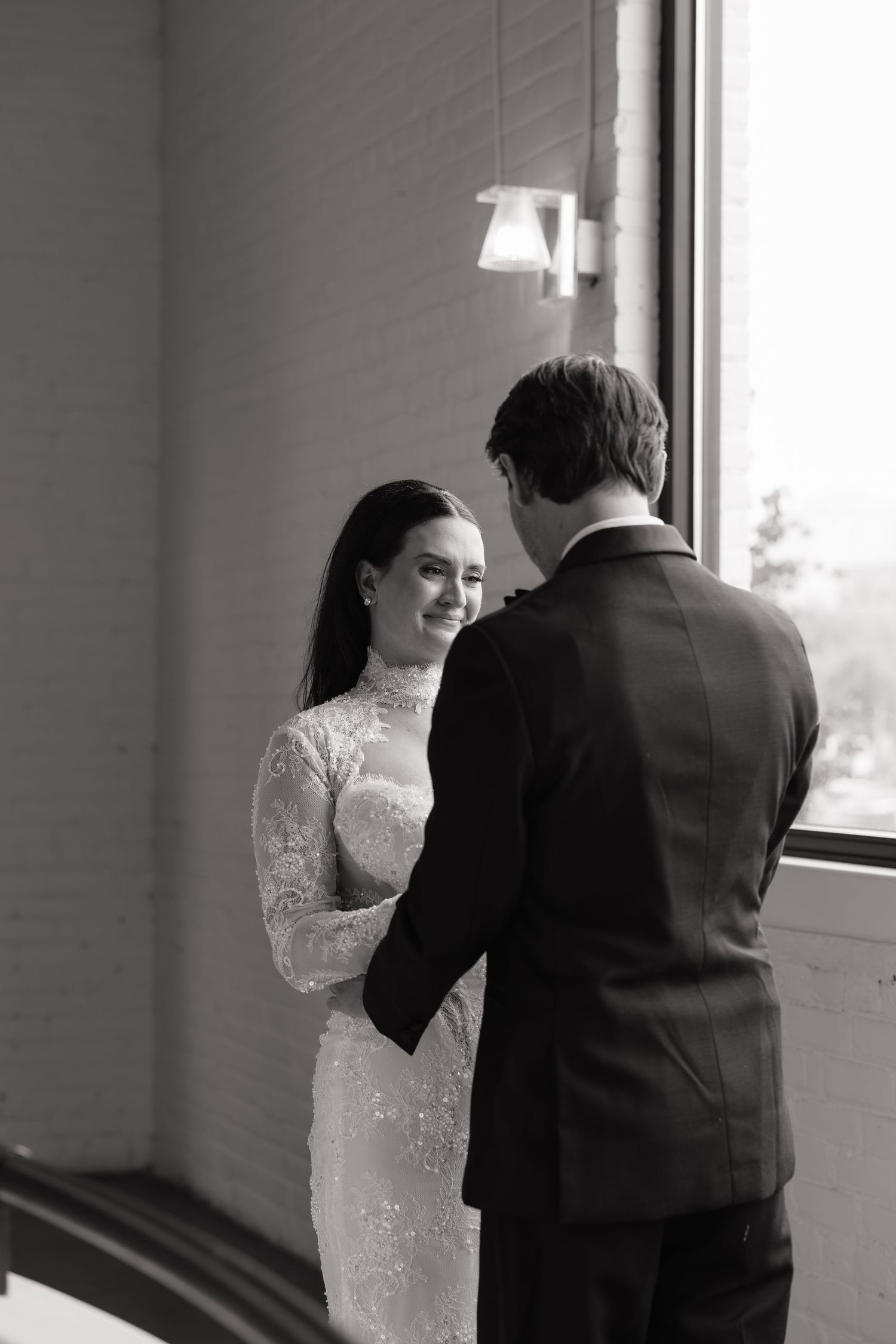 Black and white photo of a smiling bride in lace gown holding hands with groom in suit near a window.