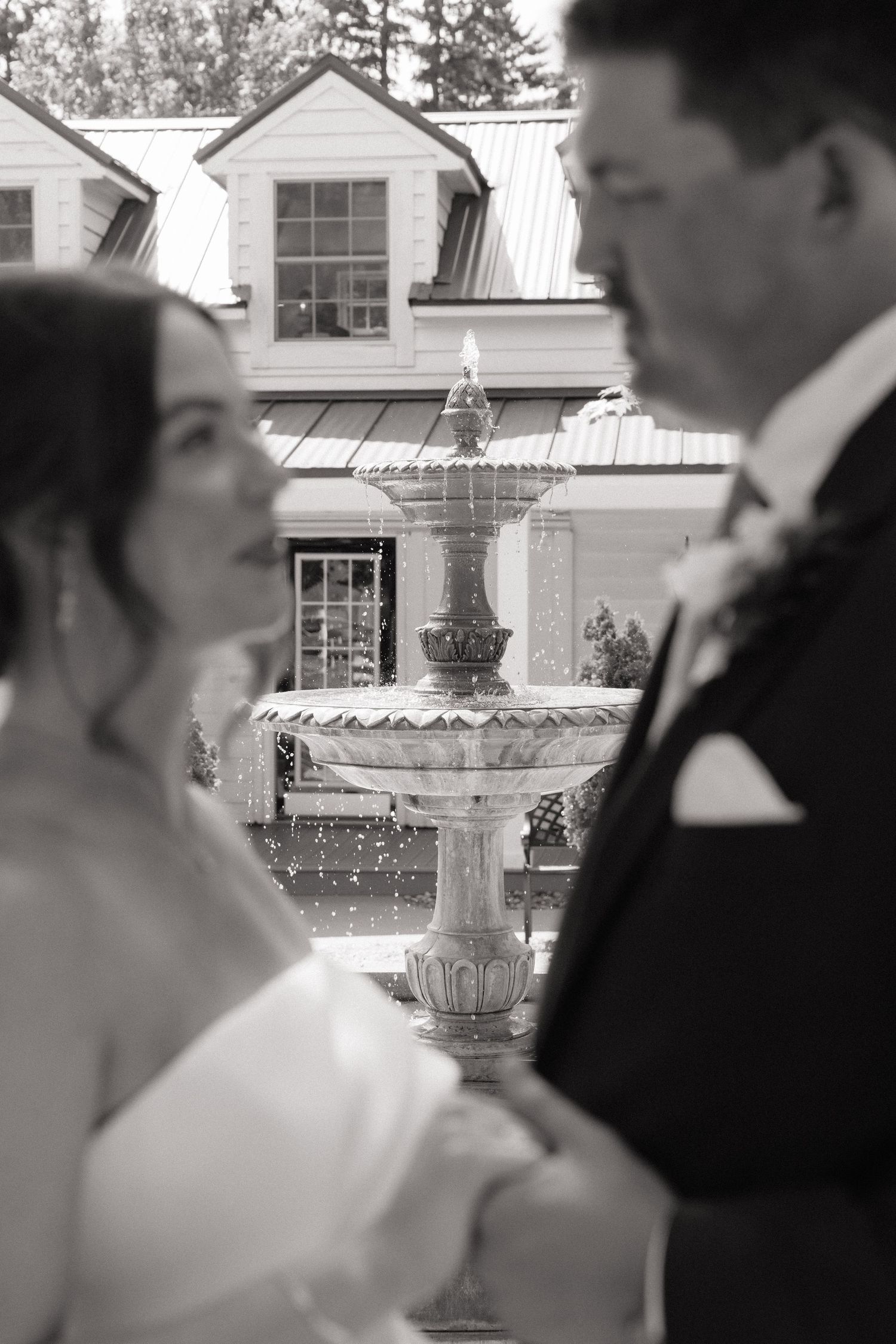 Black and white wedding photo of bride and groom with elegant fountain and venue in background.