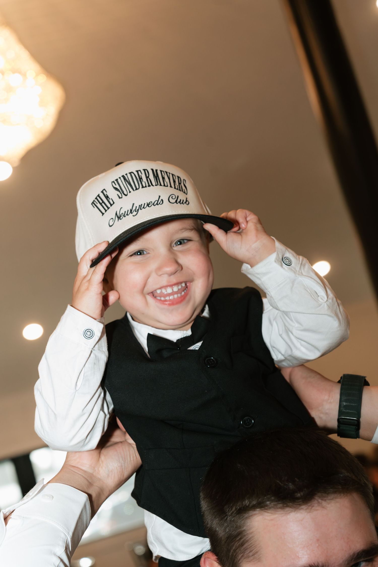 Smiling boy in black vest and bow tie wearing a The Sundermeyers Newlyweds Club hat at a wedding.