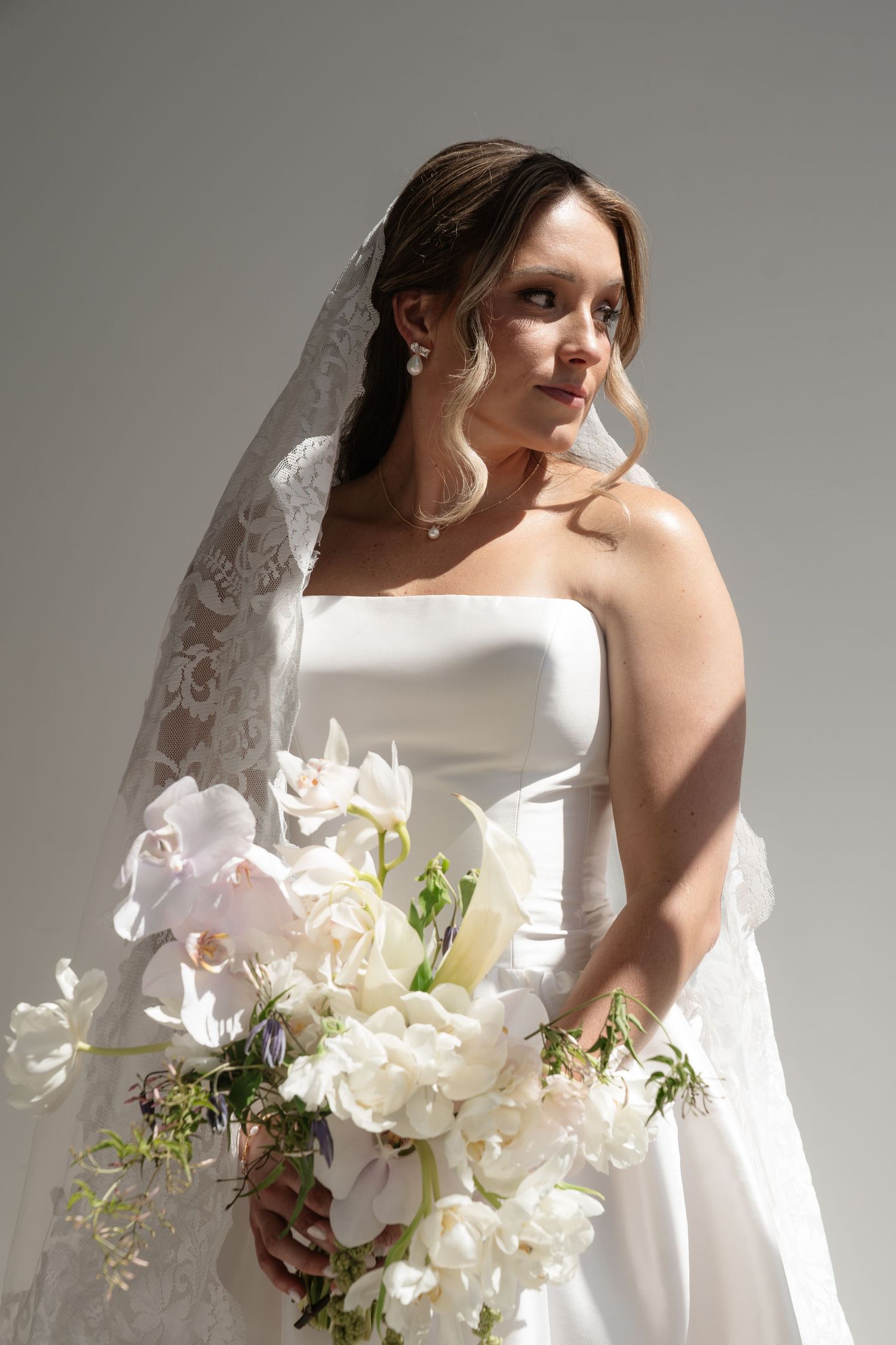 Bride in strapless white gown and lace veil holding elegant white orchid bouquet, posing gracefully against gray background.