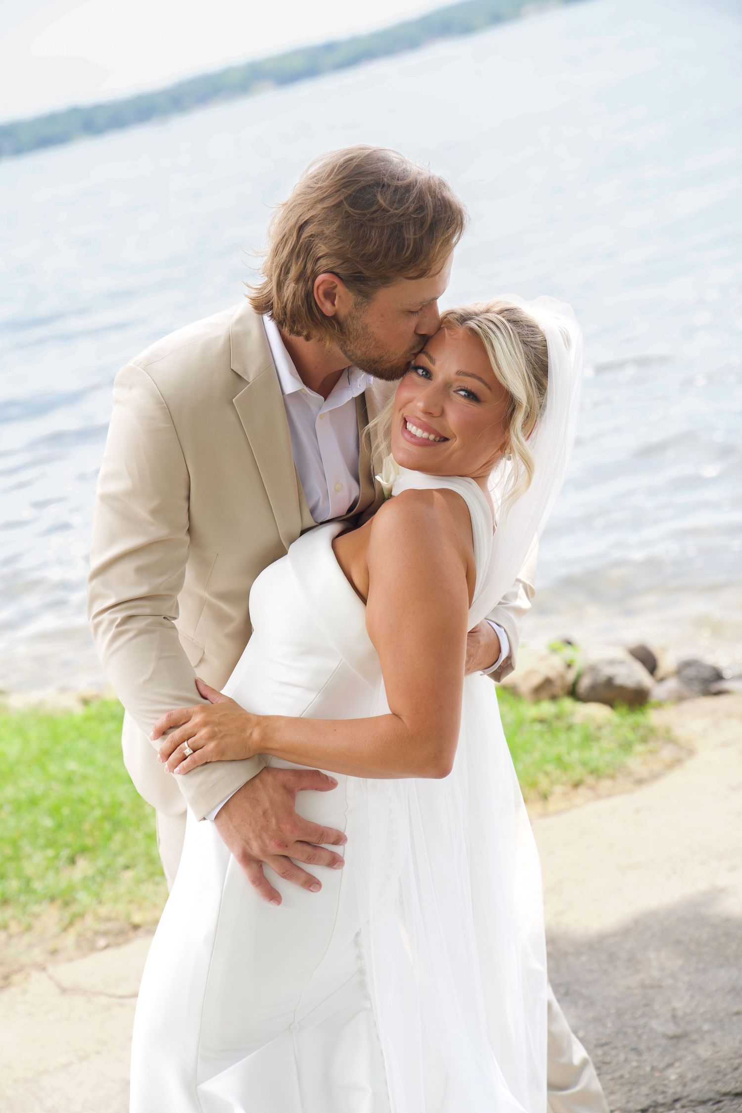 A smiling bride in a white dress embraces her groom in a tan suit by a lakeside on their wedding day.