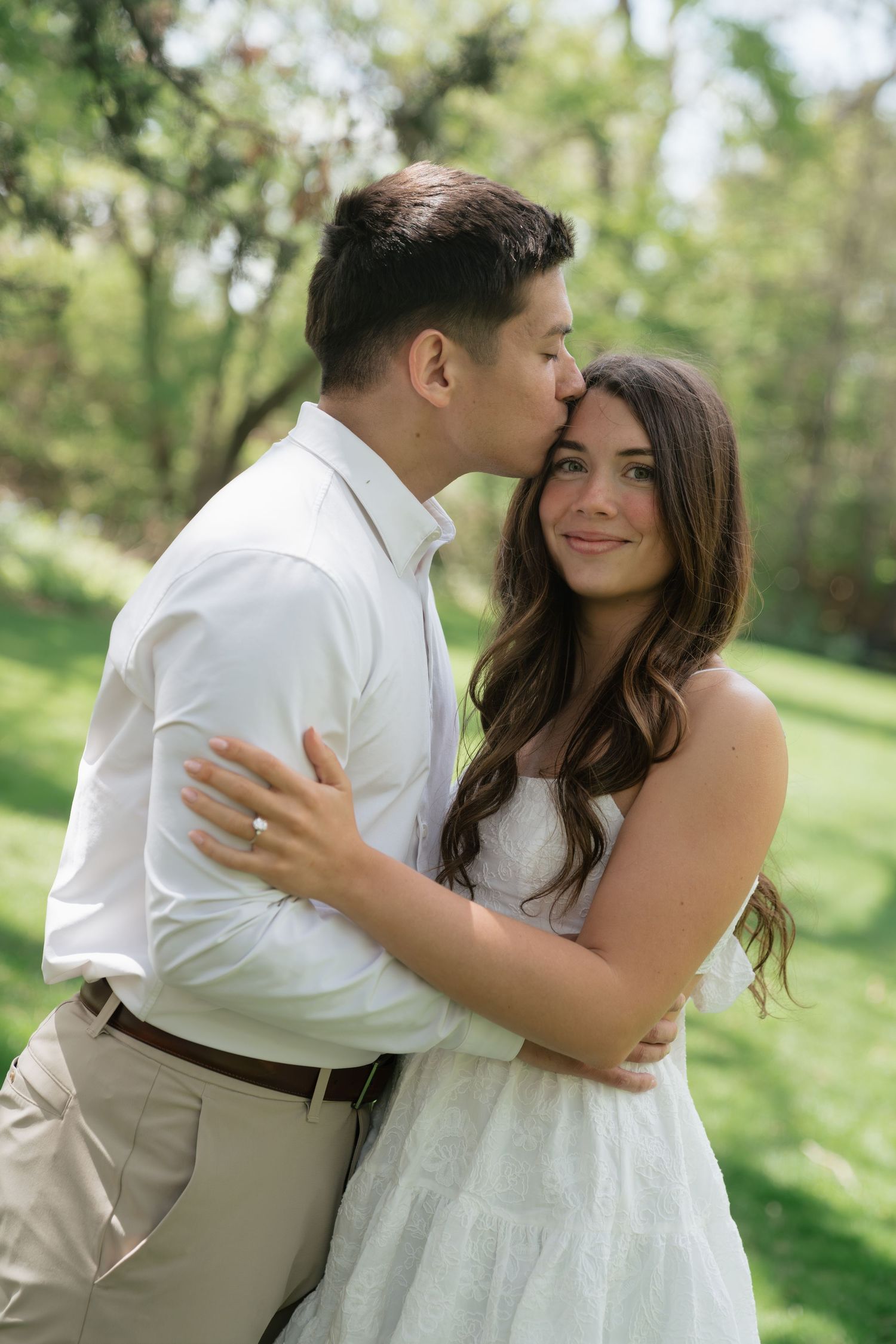 Couple embracing outdoors, man kissing woman on forehead, she smiles in white dress, lush green park background.