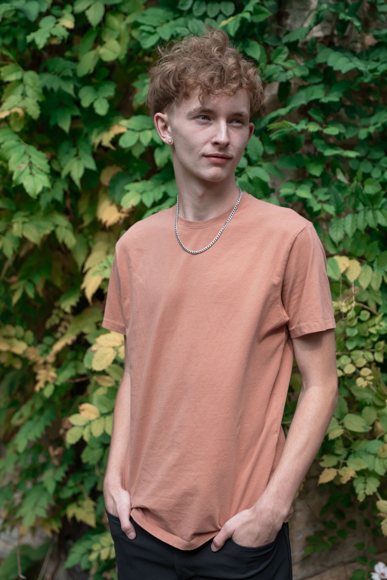Person in peach-colored t-shirt poses against ivy-covered wall in outdoor photoshoot.