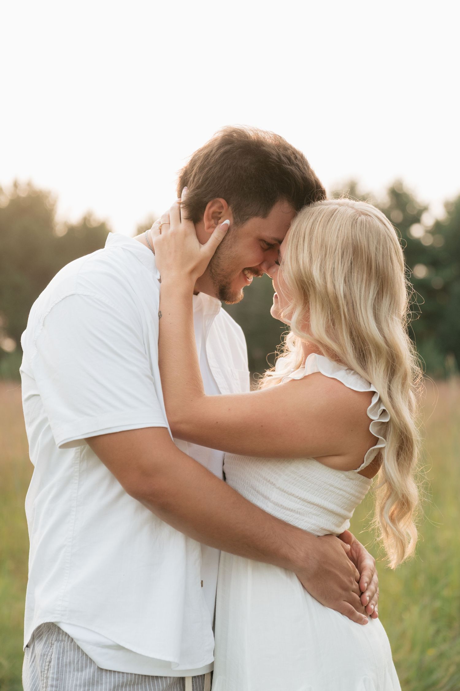 Romantic couple in white outfits embracing and smiling in a sunny green field.