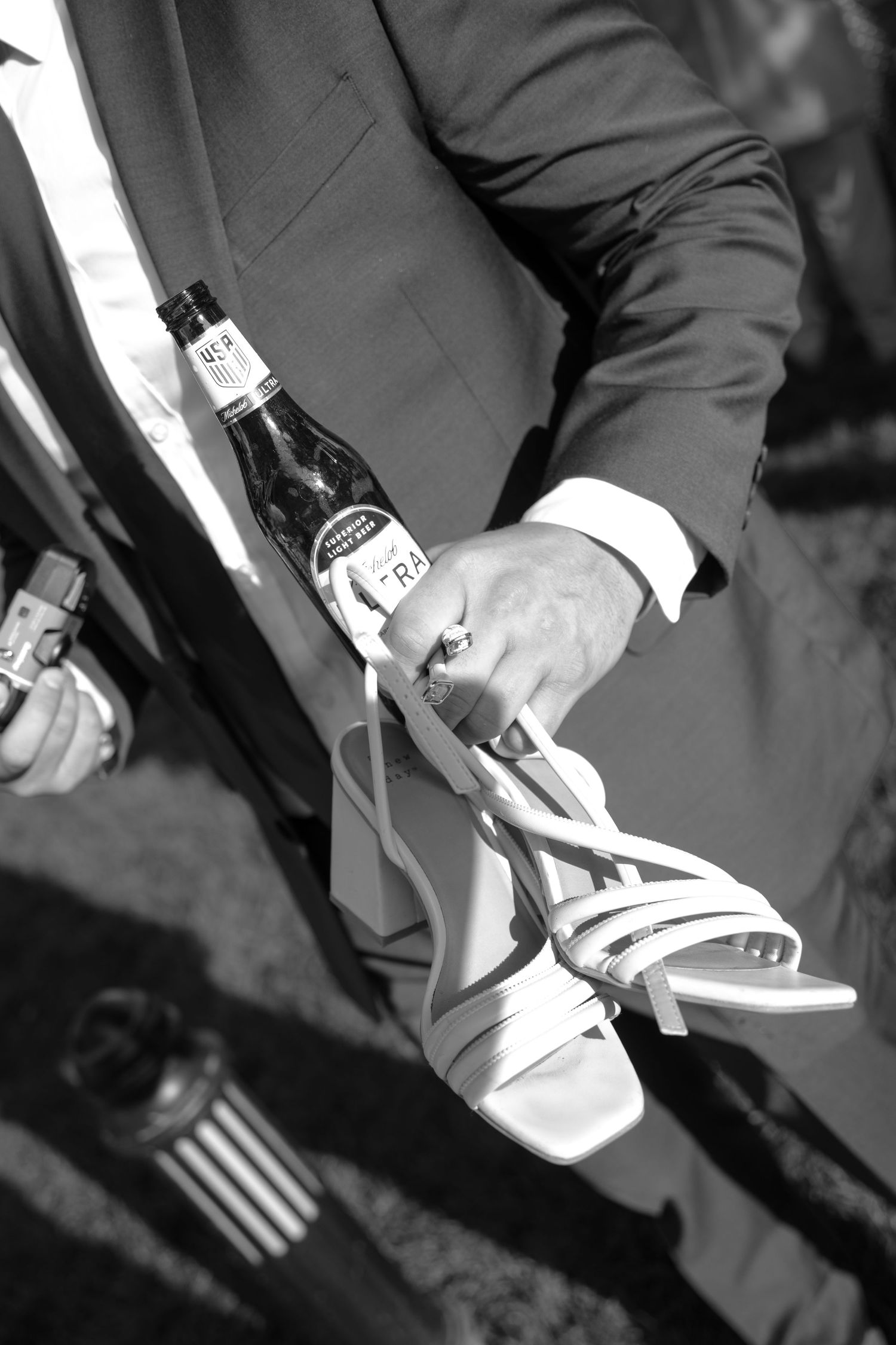 Groom holding a champagne bottle and white bridal heels in a black and white wedding detail photo.
