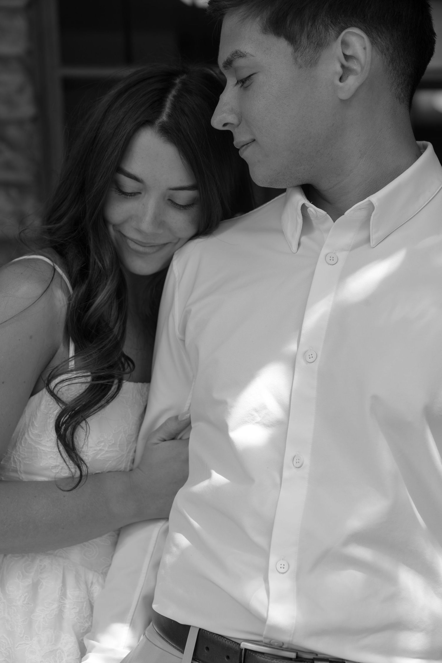 Black and white photo of a couple embracing, woman leaning on mans shoulder, both smiling softly in romantic pose.