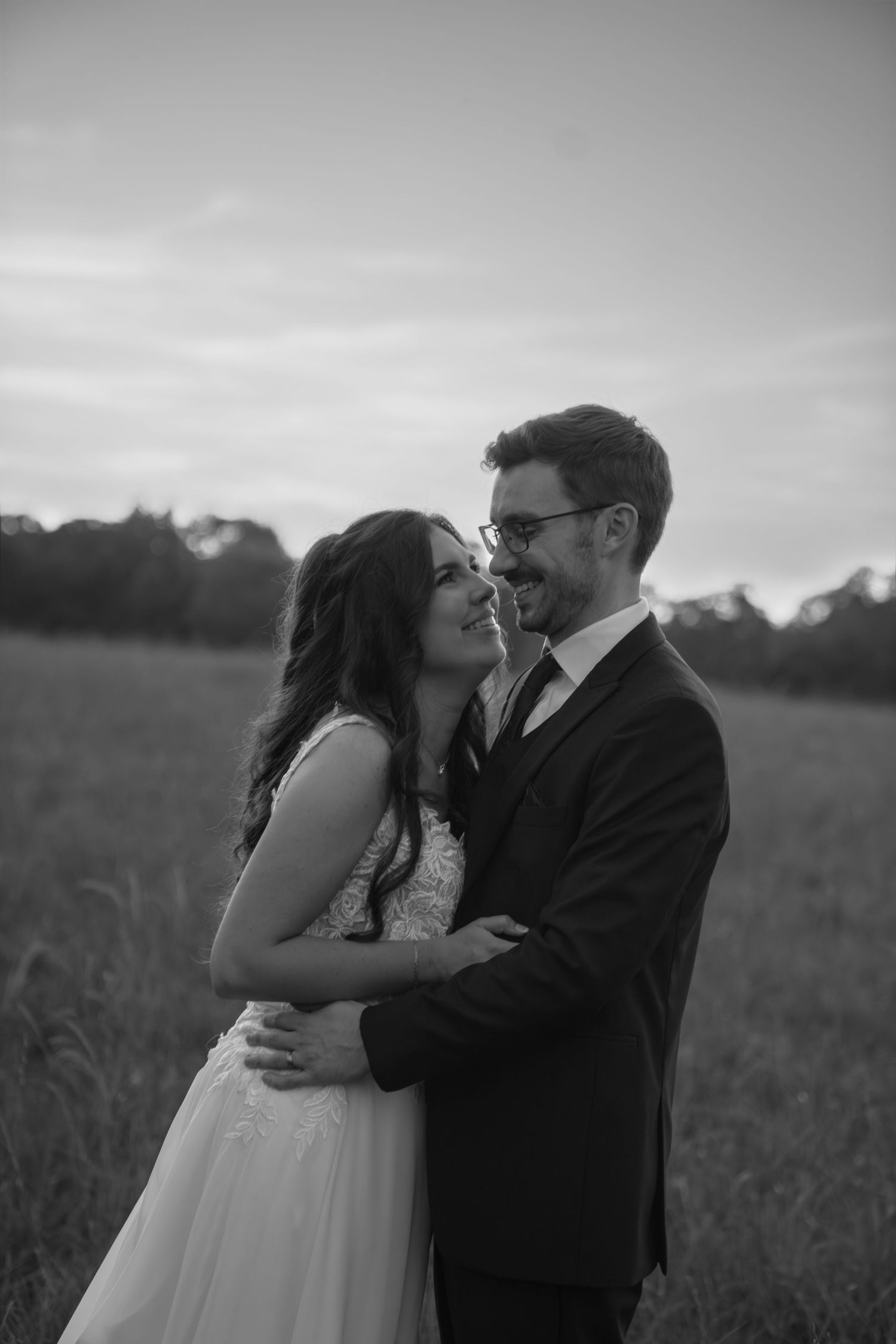 An intimate black and white portrait of a couple embracing in a misty field at dusk.