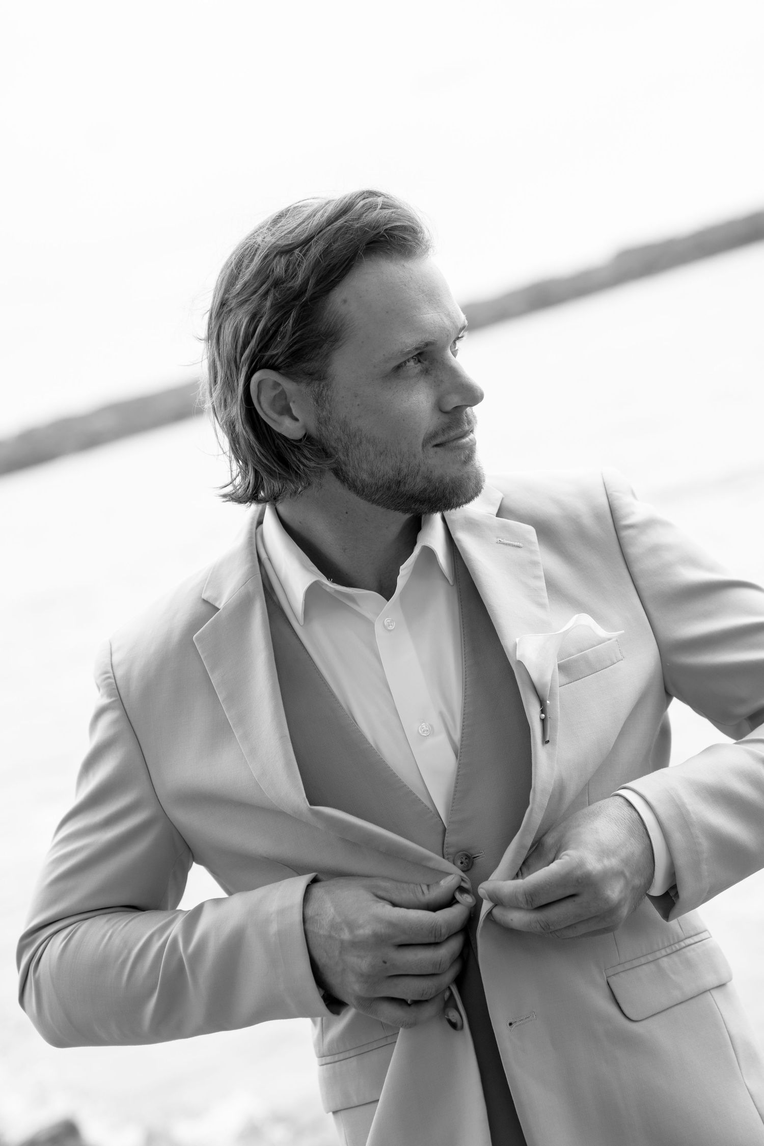 Black and white photo of a stylish man in a suit adjusting his jacket outdoors near water.