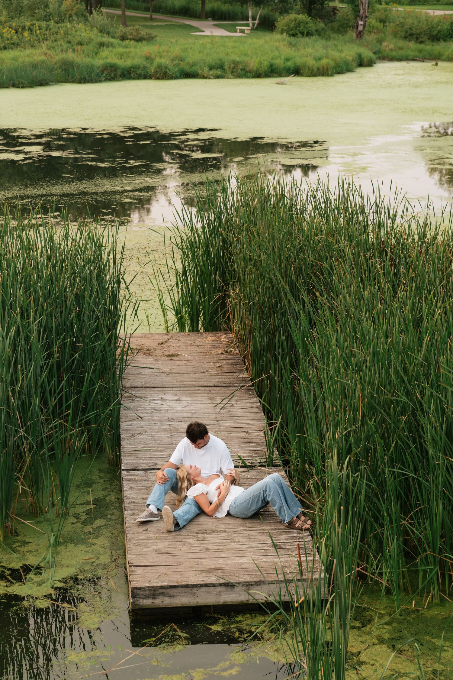 Couple relaxing on a wooden dock surrounded by tall reeds and a green pond in a lush park setting.
