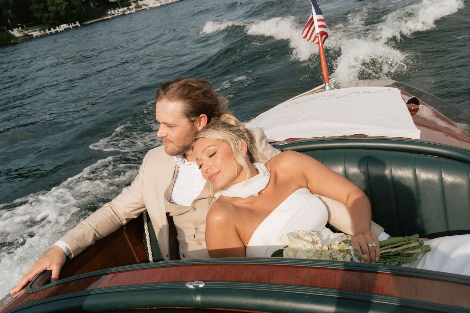 Bride and groom embracing on a classic wooden speedboat on a lake, with an American flag in the background.