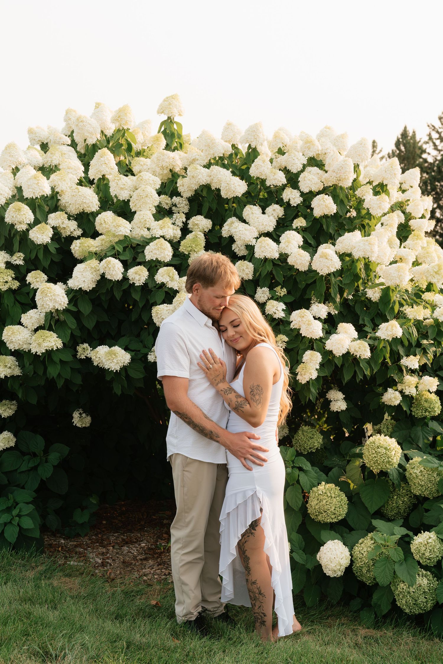Couple embracing in front of lush white hydrangea bushes, woman in white dress with tattoos, man in white shirt.