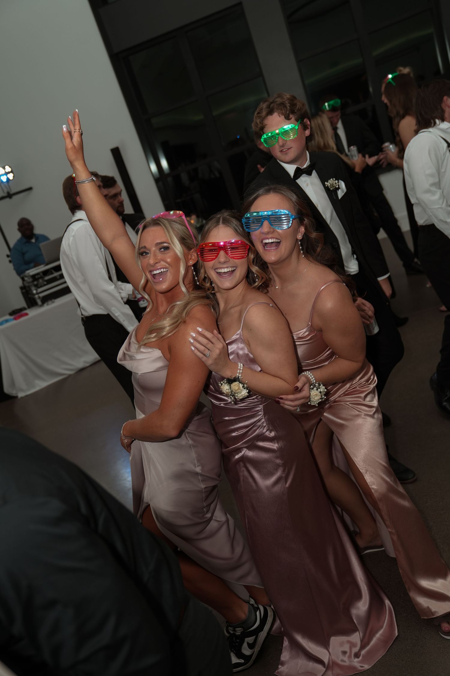Wedding party guests in brown satin dresses dance and pose with colorful sunglasses on the dance floor.