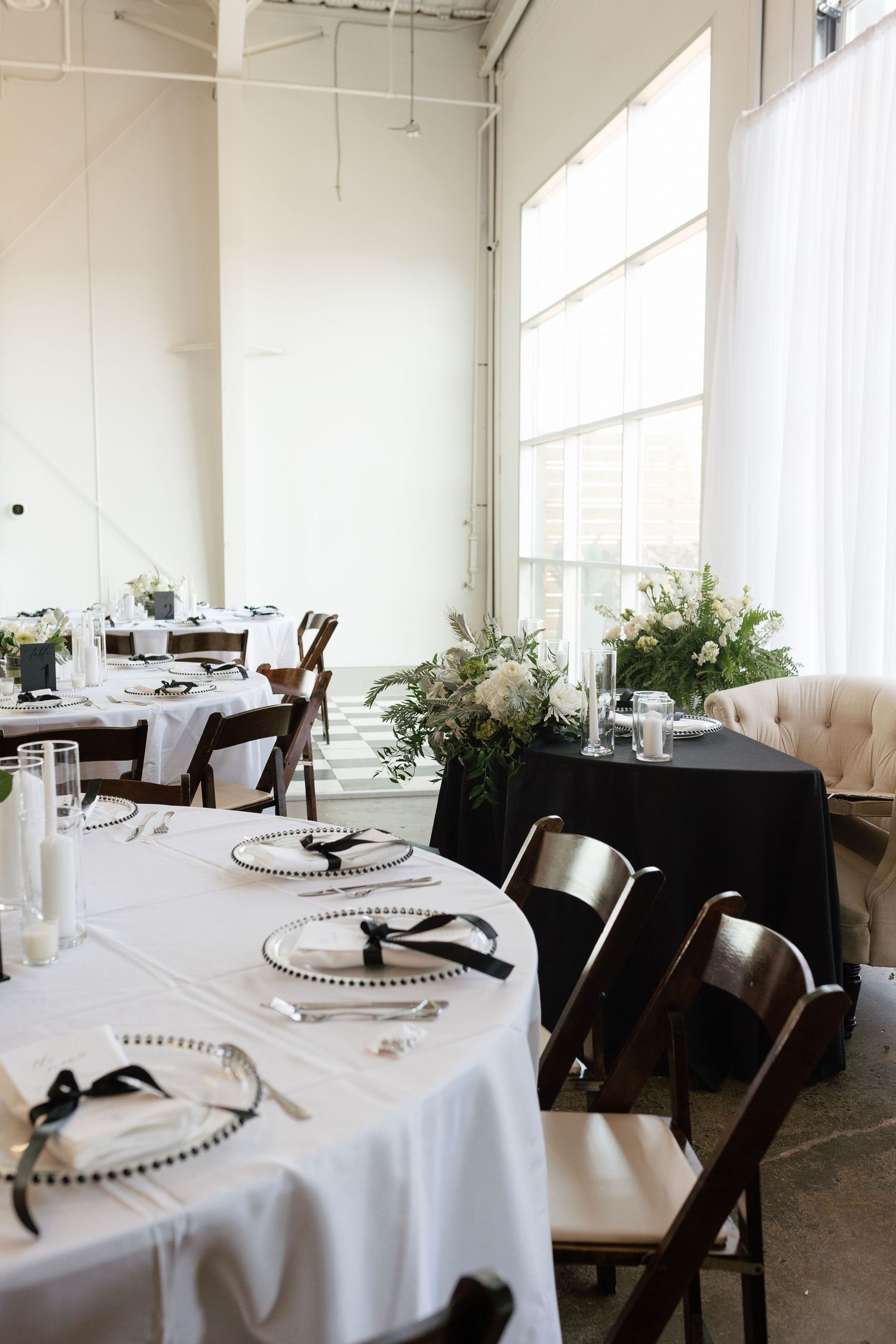 Elegant wedding reception tables with white linens, black ribbon accents, charger plates, and lush floral centerpieces in a bright venue.