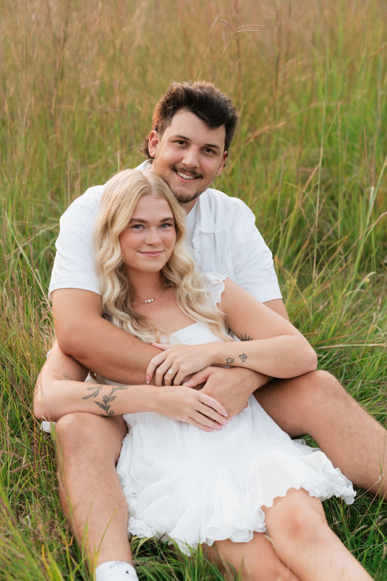 A smiling couple in white outfits sitting together in a lush green field, sharing a warm embrace.