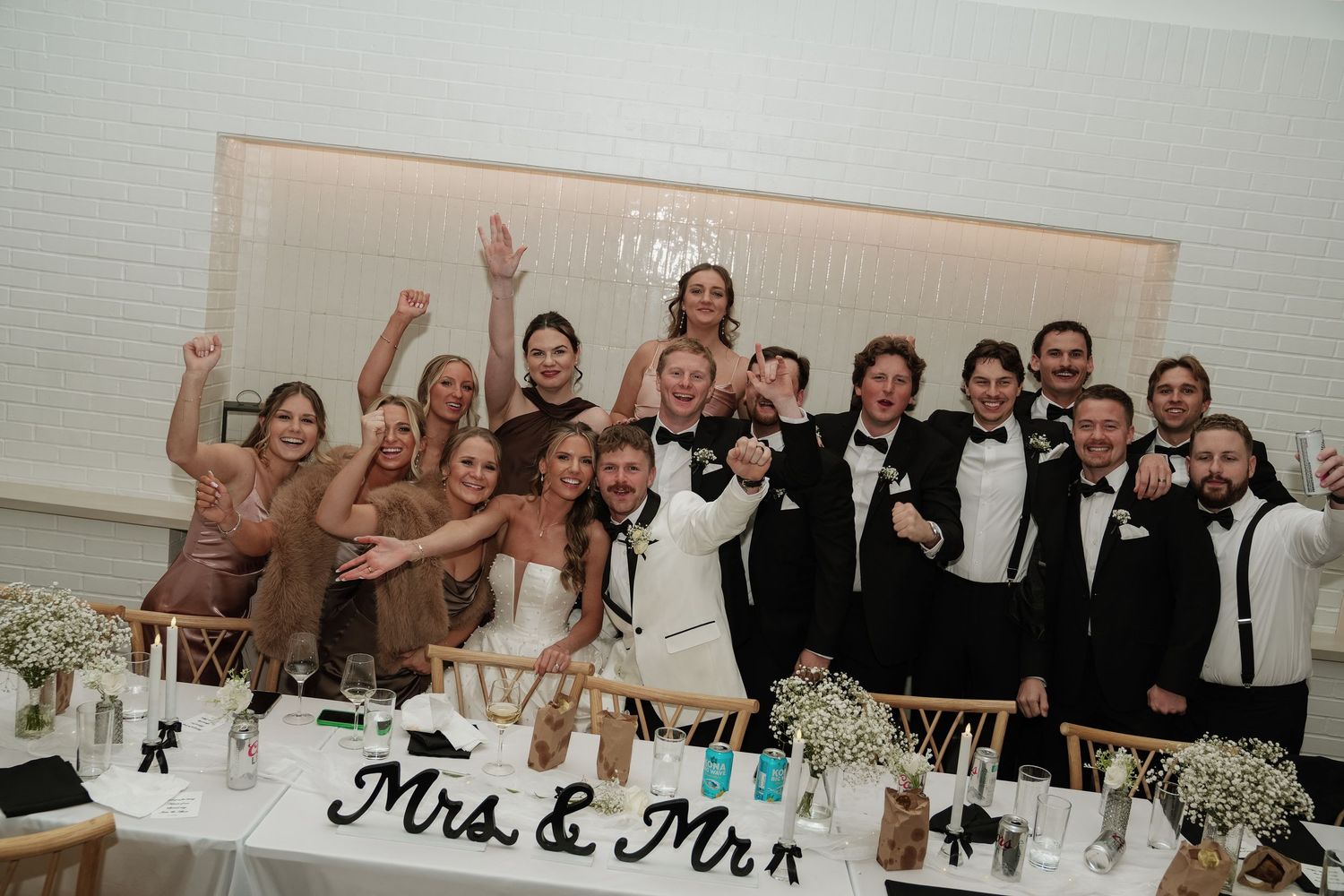 Wedding reception guests raise their glasses in celebration at a long banquet table decorated with white floral centerpieces.