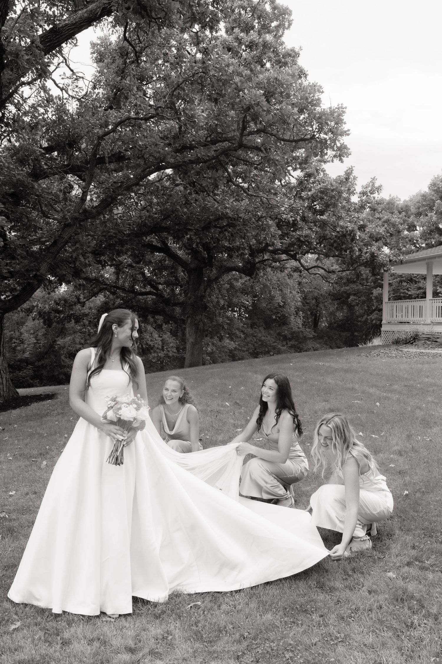 Black and white photo of a bride holding bouquet while bridesmaids adjust her wedding gown train outdoors.
