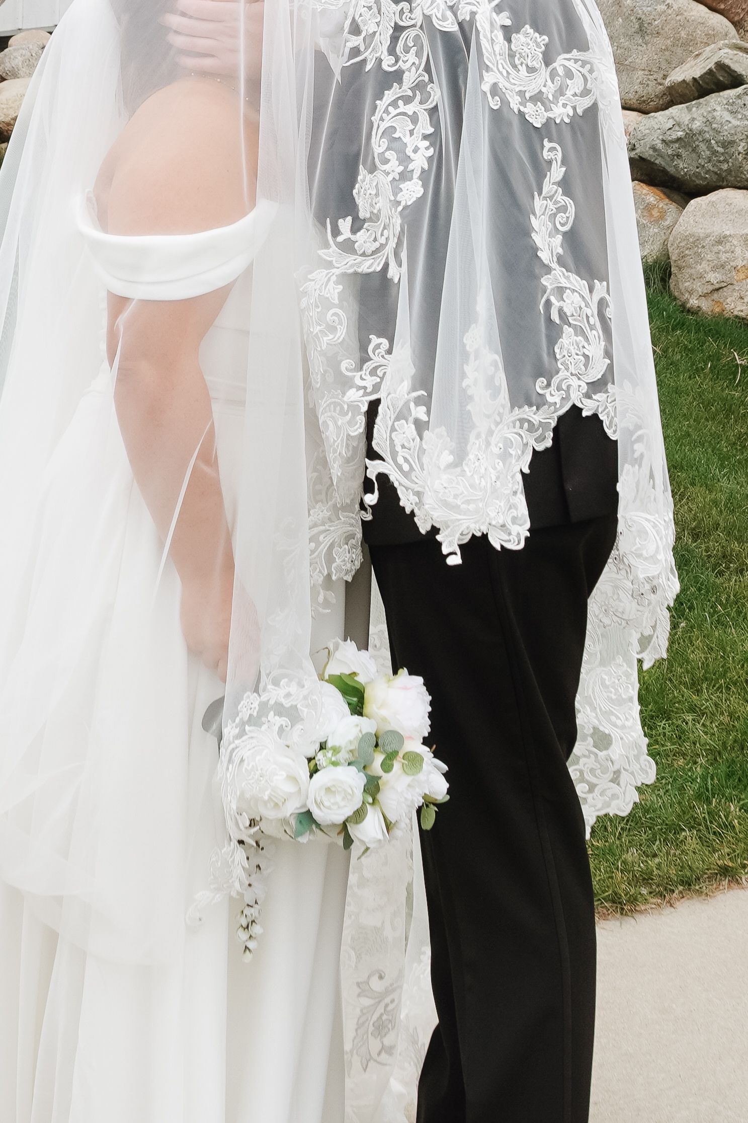 Bride and groom standing together outdoors, bride holding white rose bouquet, wearing lace-trimmed veil and white off-shoulder gown.