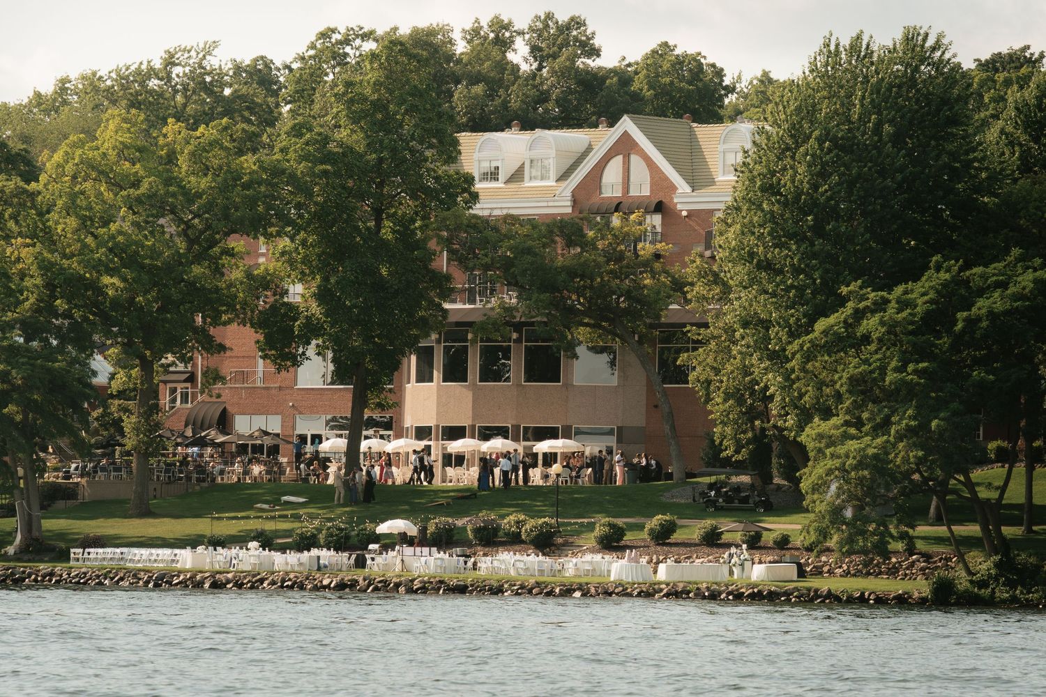 Waterfront venue with brick building surrounded by lush trees, guests gathered on lawn for outdoor event near calm lake.