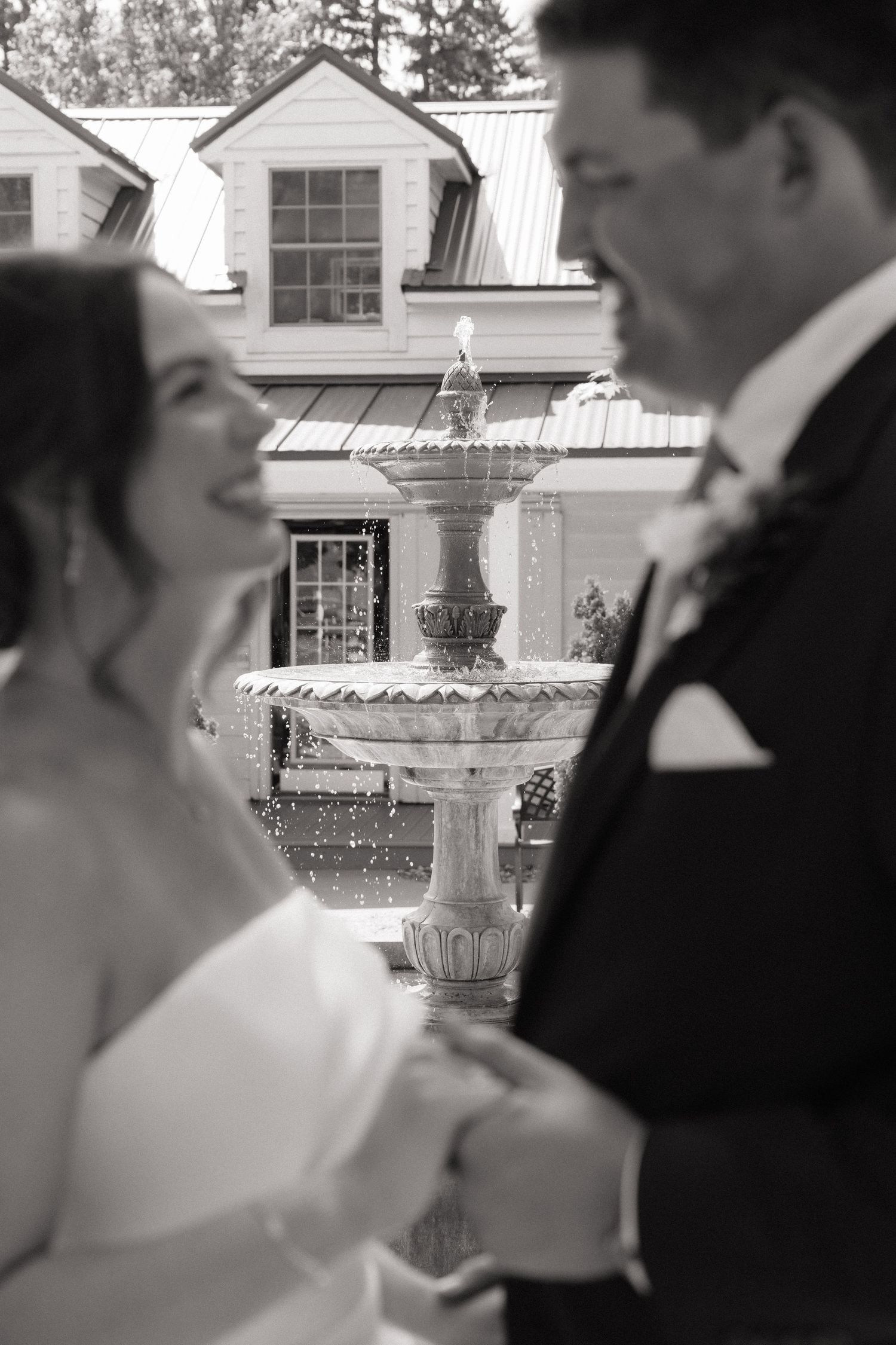 Black and white wedding photo of smiling bride and groom holding hands in front of elegant fountain.