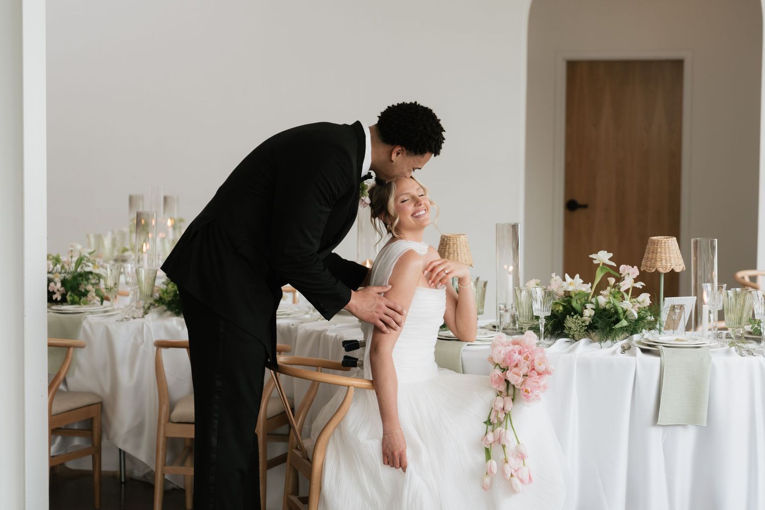 Groom in black suit kissing bride in white dress on forehead at elegantly decorated wedding reception table.