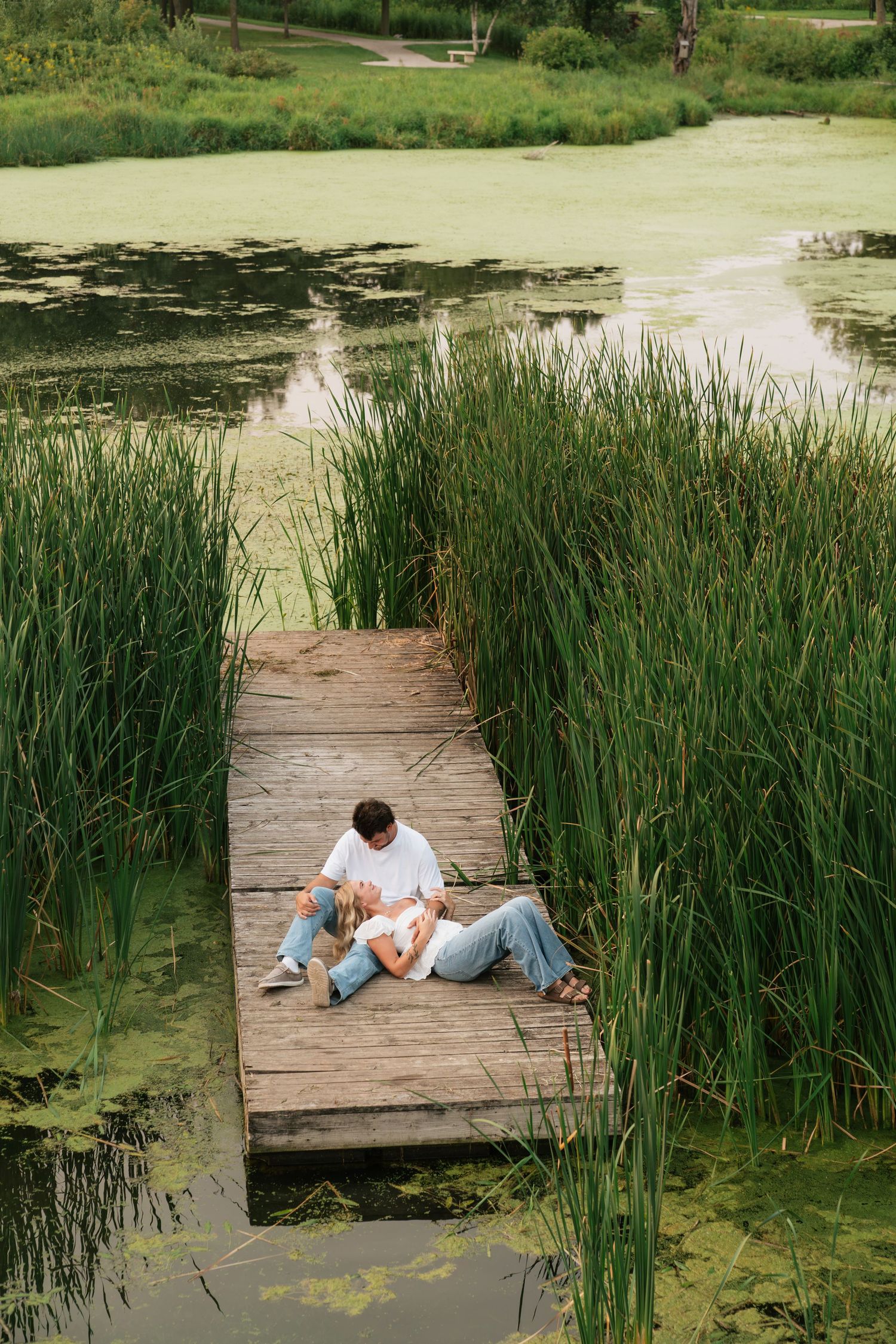 Couple relaxing on a wooden dock surrounded by tall reeds and a green algae-covered pond in a lush park.