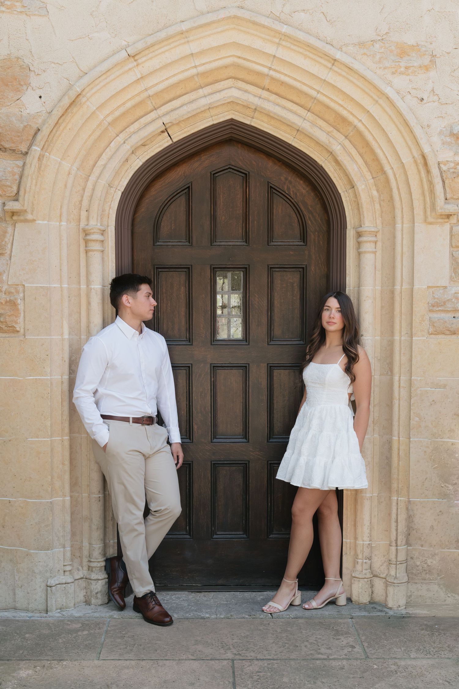 Couple posing in front of ornate arched wooden door at stone building, man in white shirt and woman in white dress.