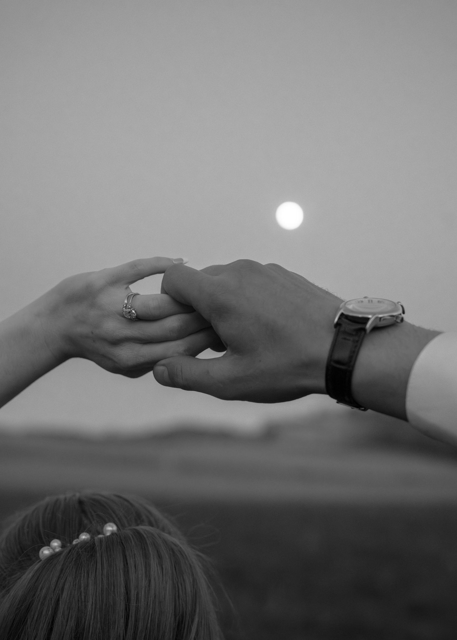 Black and white photo of couple holding hands with moon in background, woman wearing ring, man wearing watch.