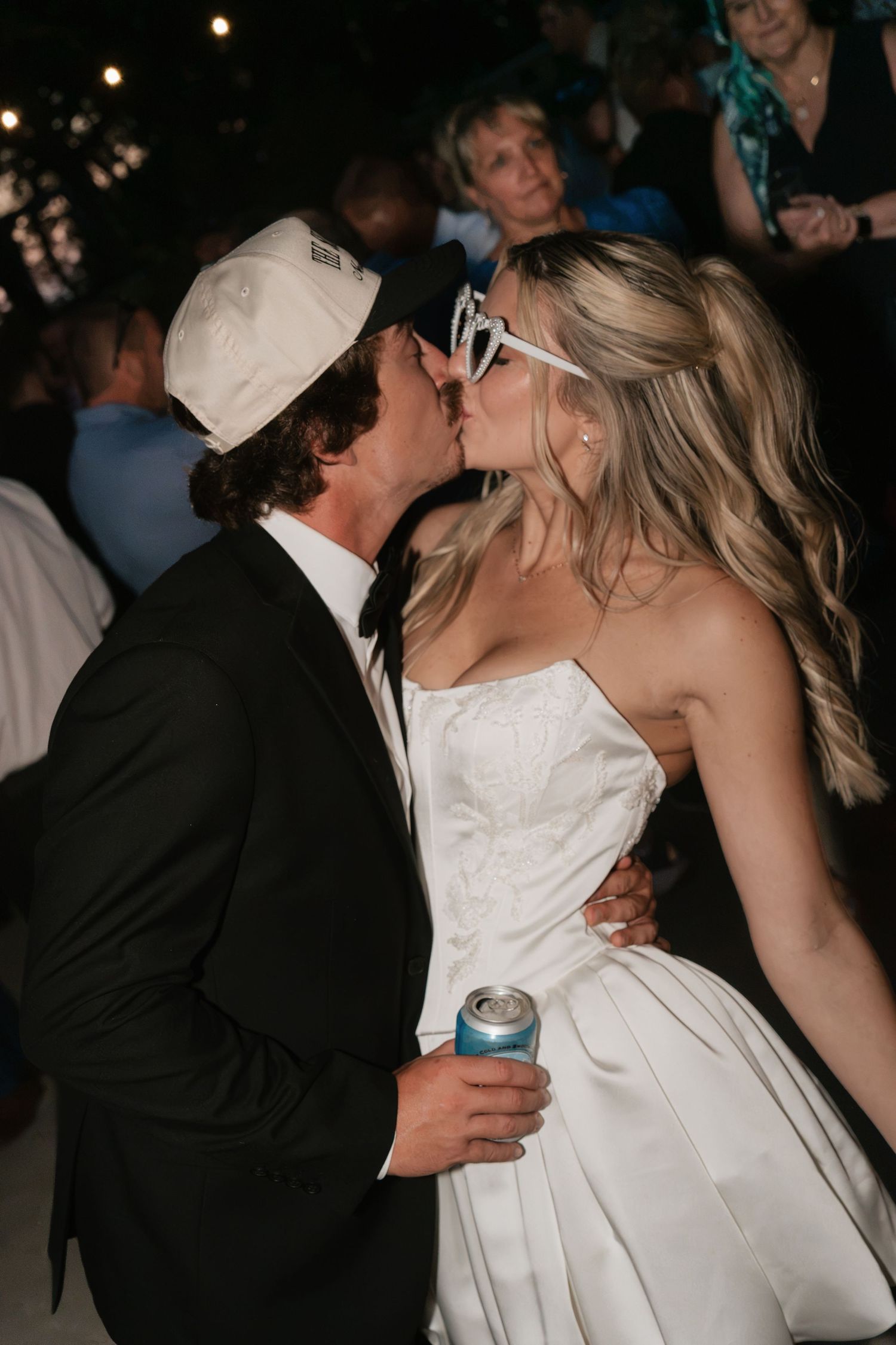 A bride and groom share a kiss at their wedding reception, she in a white gown with sunglasses, he in a suit with a cap.