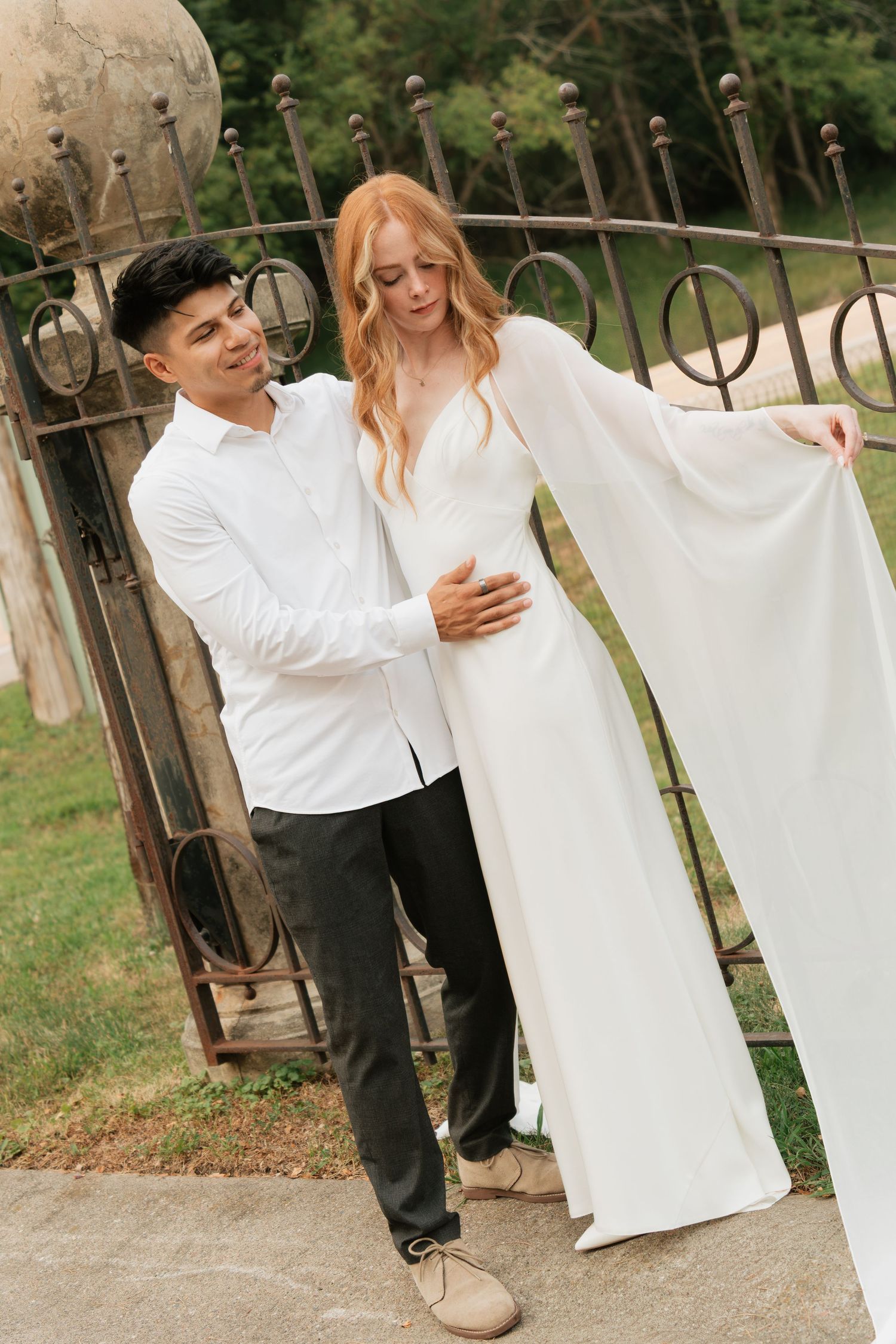 A groom in white shirt and dark pants stands with a bride in a flowing white gown near ornate iron gates outdoors.