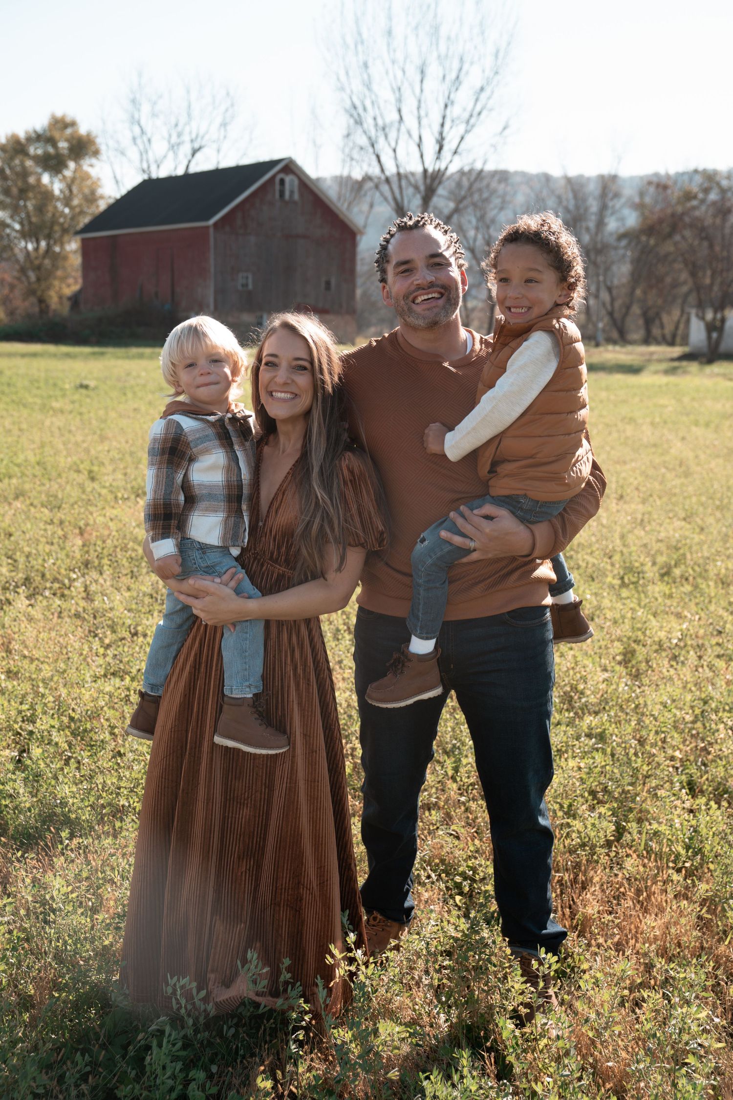 A family of four poses together in front of a red barn during golden hour.