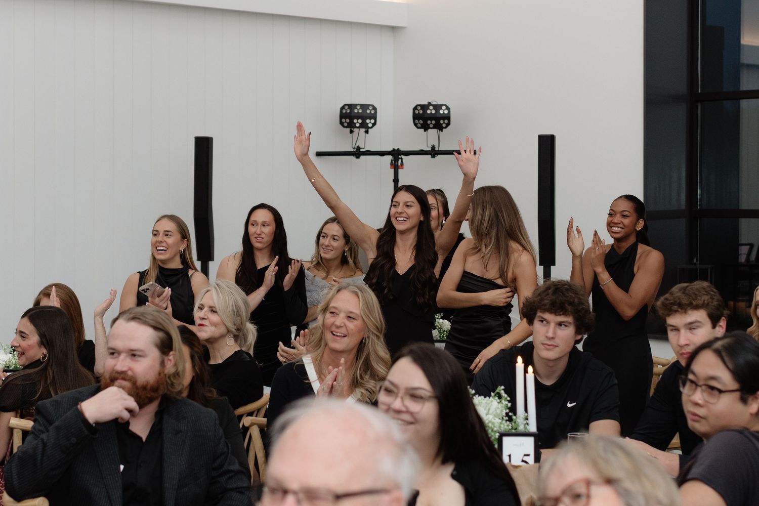 A group of people in black attire sitting and raising their hands during an indoor event.