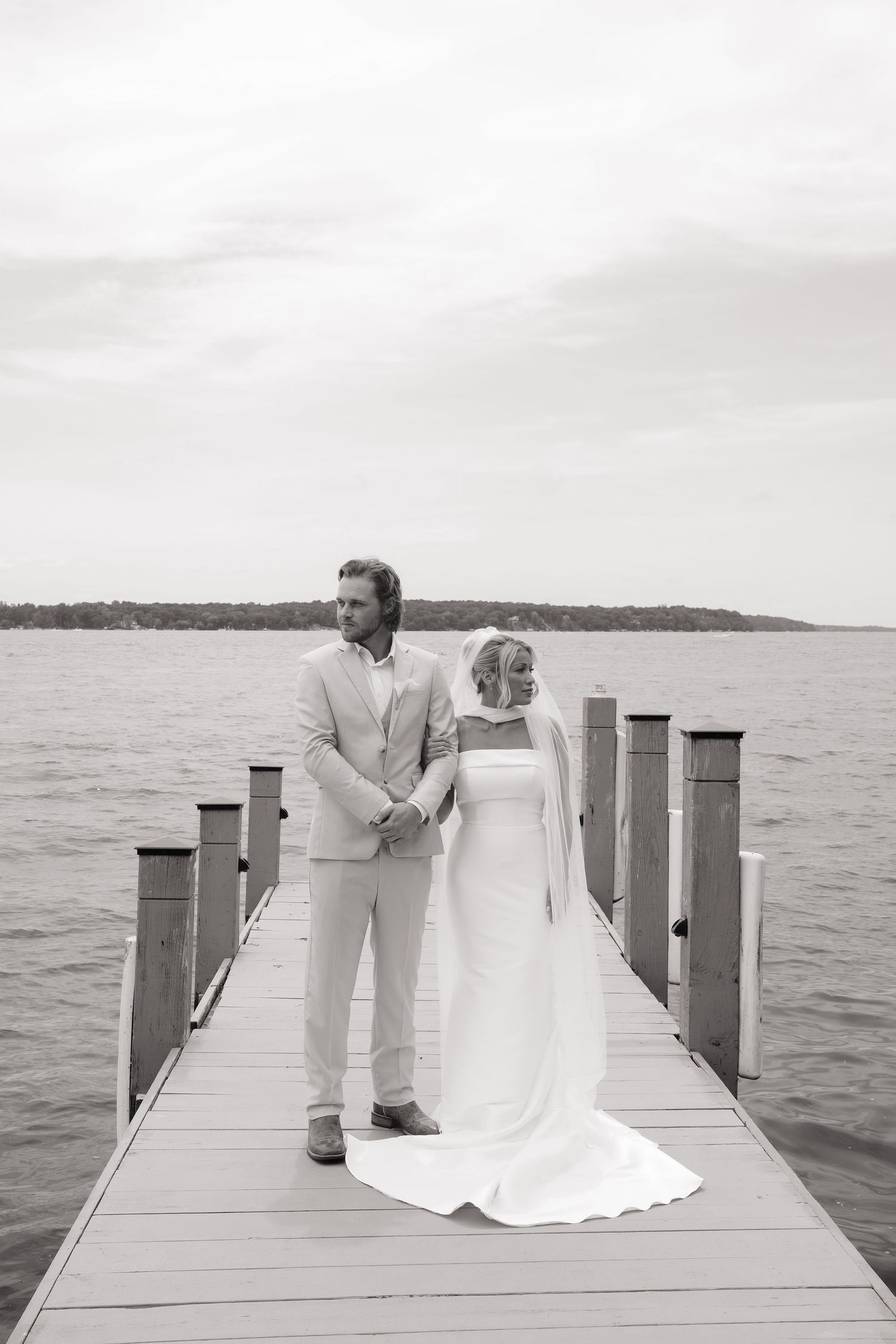 Black and white photo of a bride and groom standing on a dock overlooking a serene lake on their wedding day.