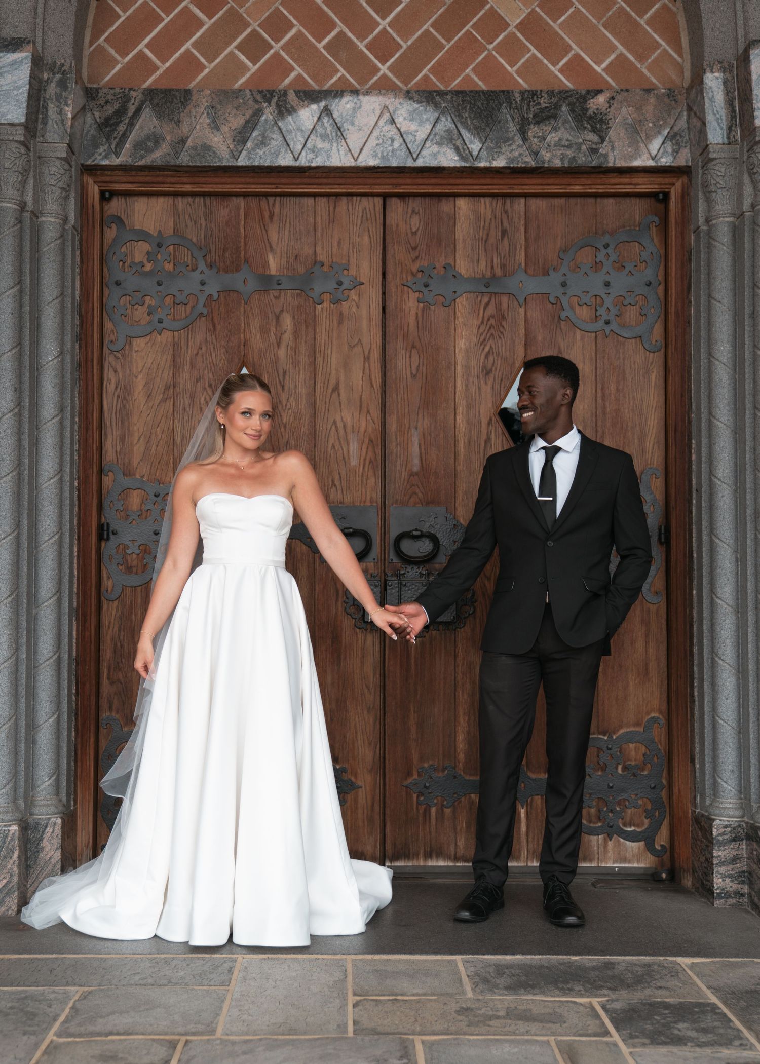 A bride in a white gown and groom in a black suit hold hands before ornate wooden doors at their wedding.