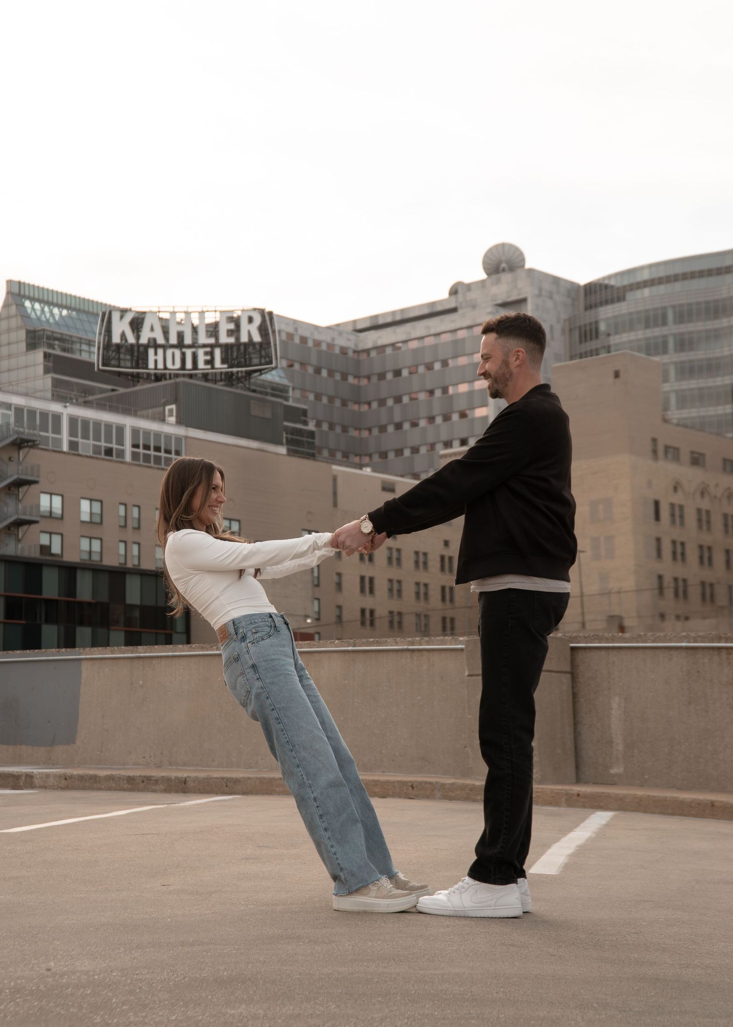 A couple holds hands and dances on a rooftop parking lot with the Kahler Hotel building in the background.