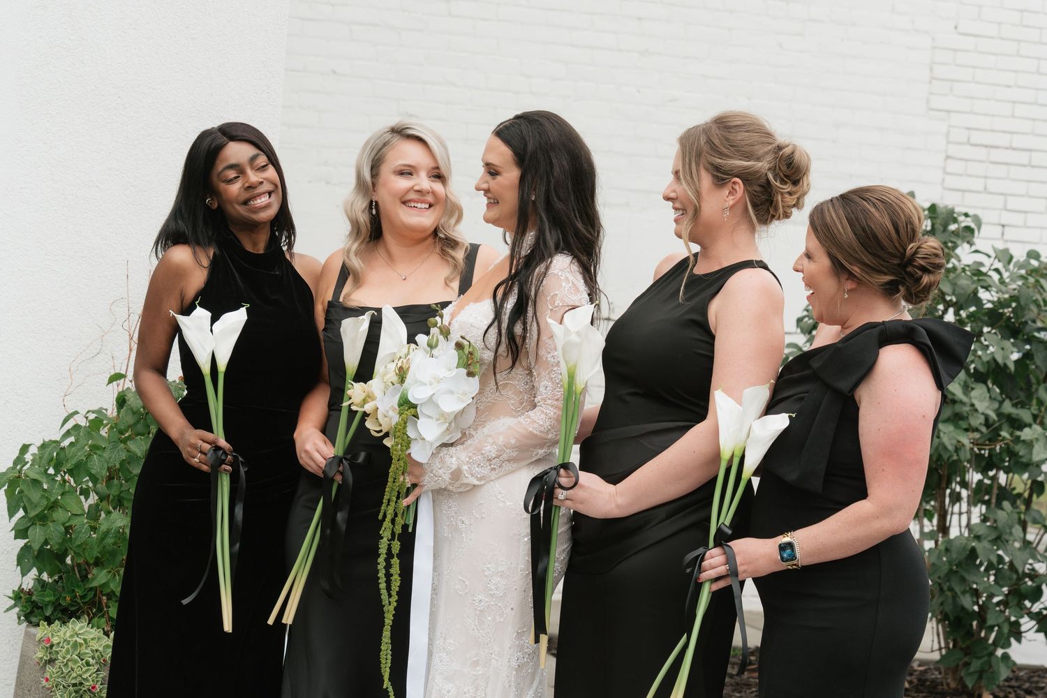Bride in white lace gown with four bridesmaids in black dresses holding white calla lilies, smiling outdoors.
