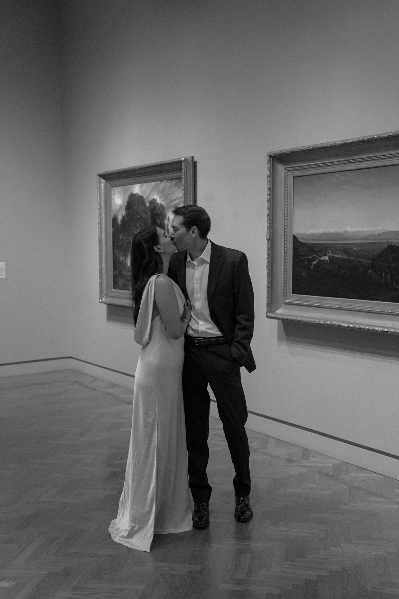 A couple sharing an intimate moment in an art gallery between framed landscape paintings.