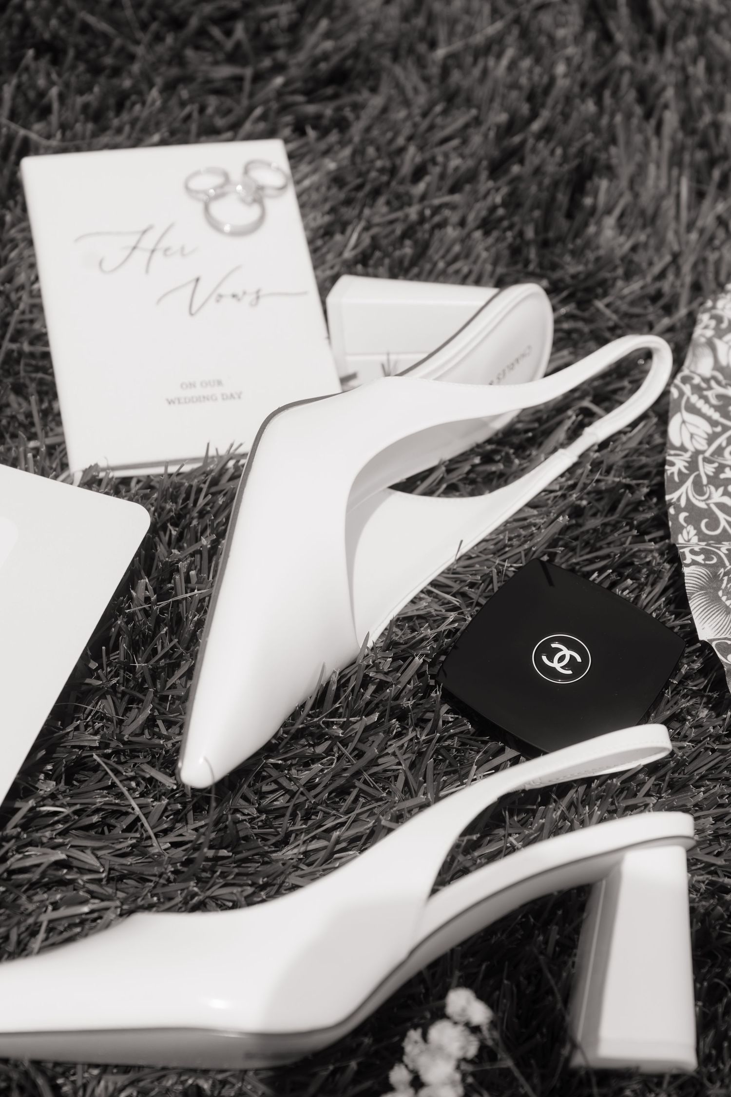 Black and white photo of white bridal heels, Chanel compact, and wedding vows book on grass.