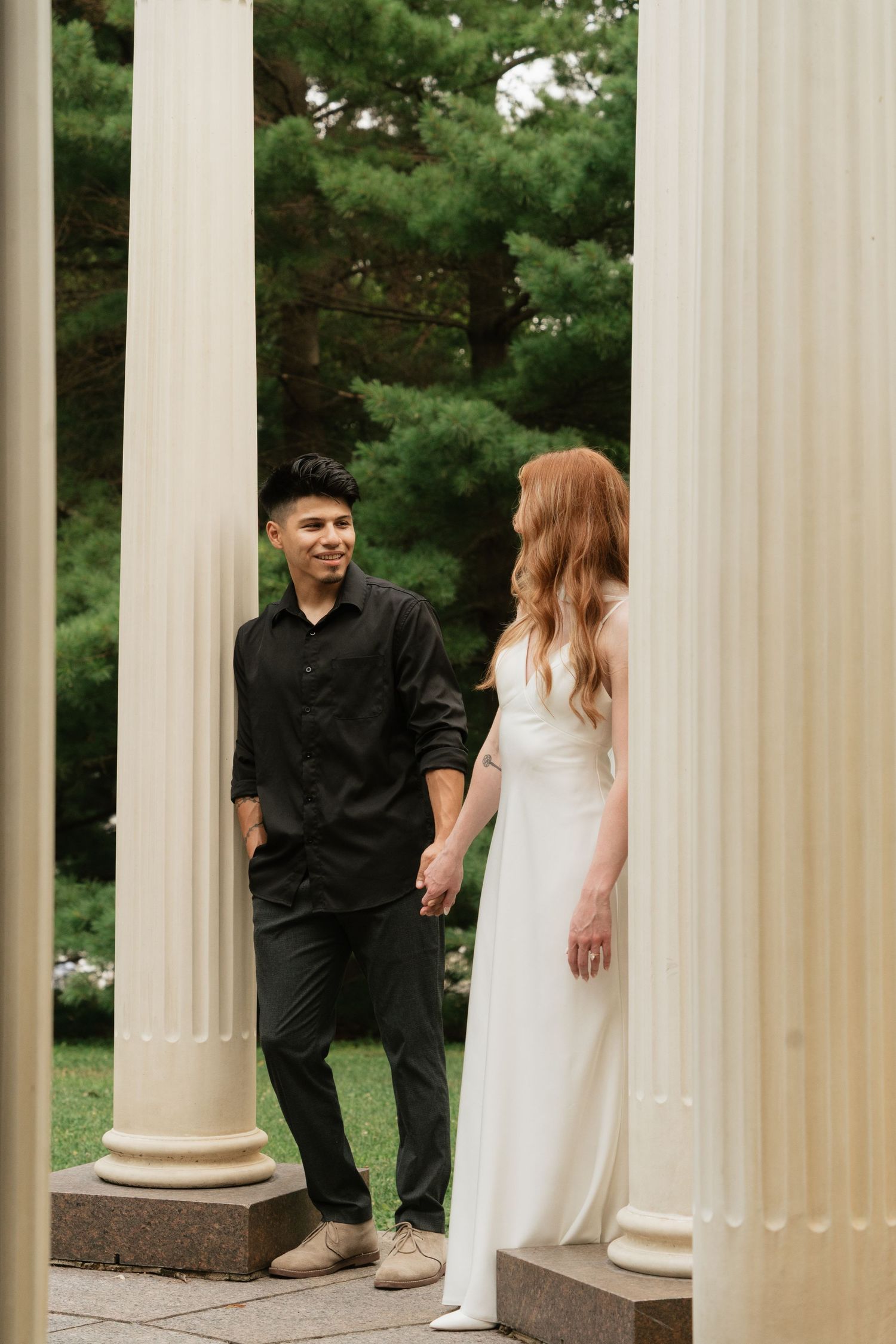 Couple holding hands between white columns, man in black outfit and woman in white wedding dress, lush green trees background.