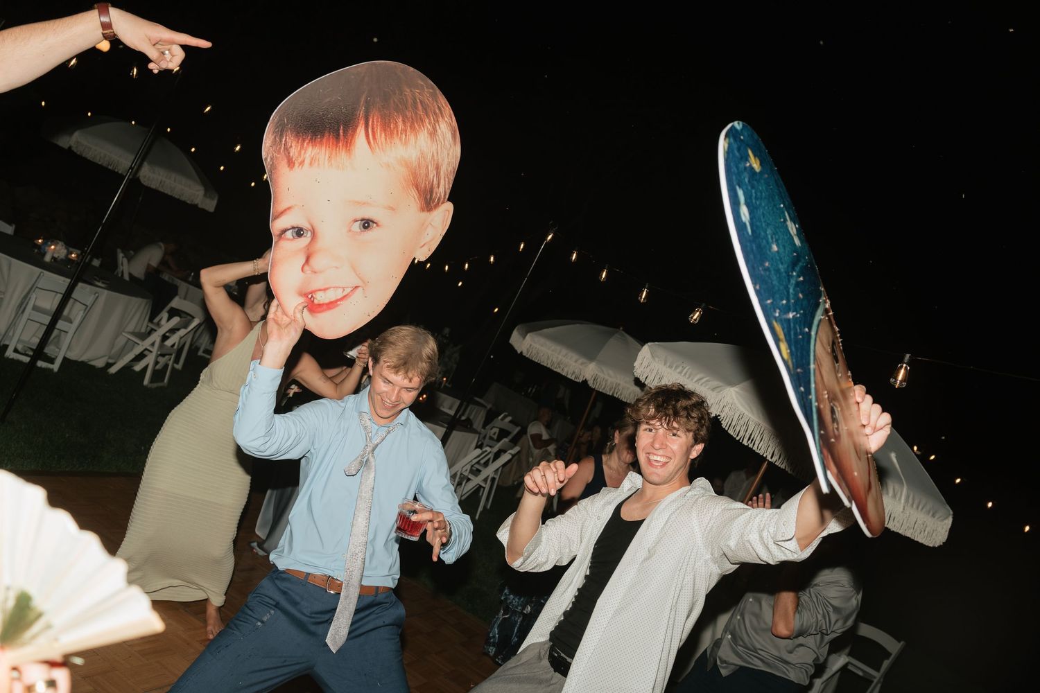 Young people dancing at a nighttime outdoor event, one holding a large child face cutout and another with a decorative fan.